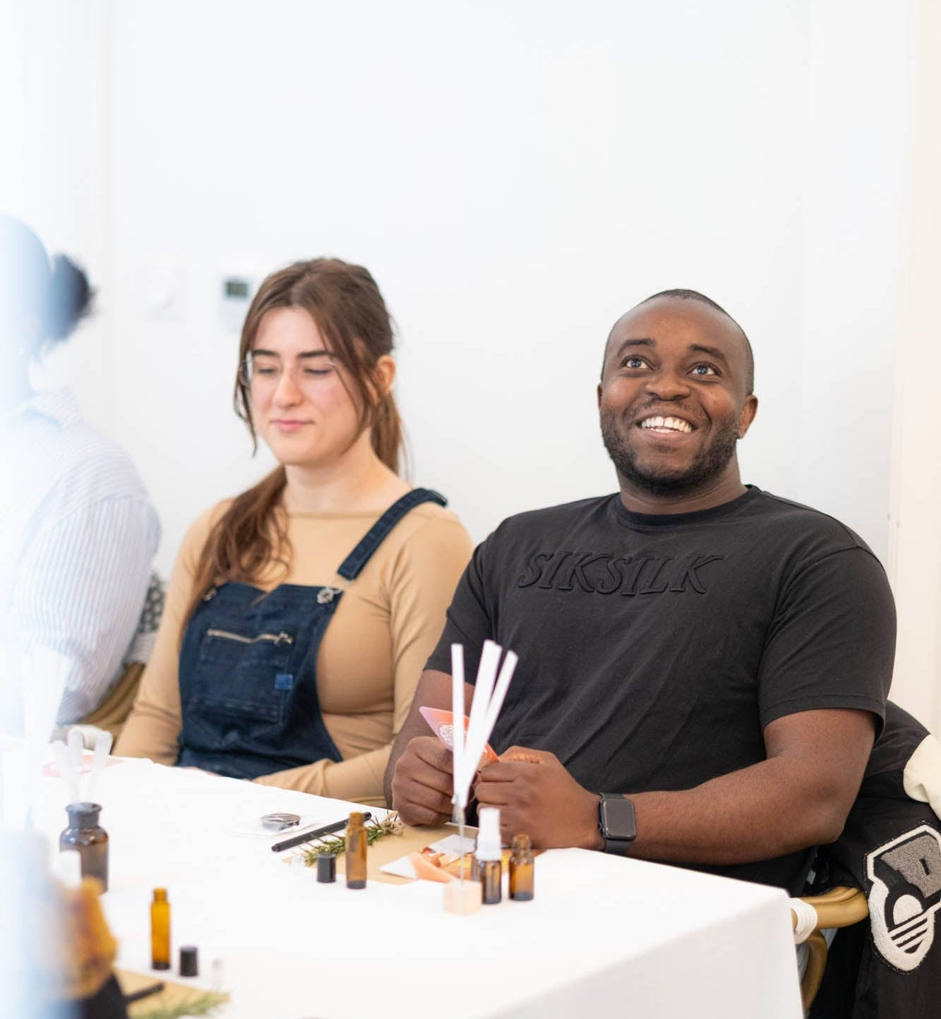 A smiling man in a black t-shirt sits at a white table holding reed diffusers, with a woman in denim overalls beside him. Small amber bottles and diffuser components are arranged on the table in a bright indoor setting.