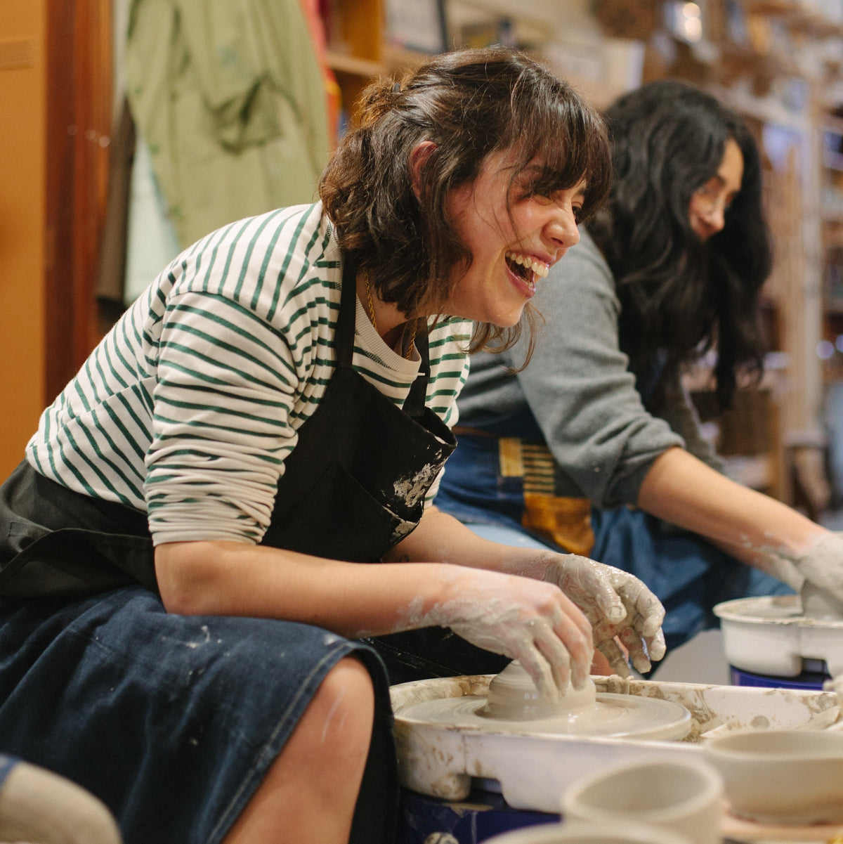 A woman in a pottery studio laughing while shaping clay on a wheel, wearing a striped shirt and black apron, with another person working in the background.