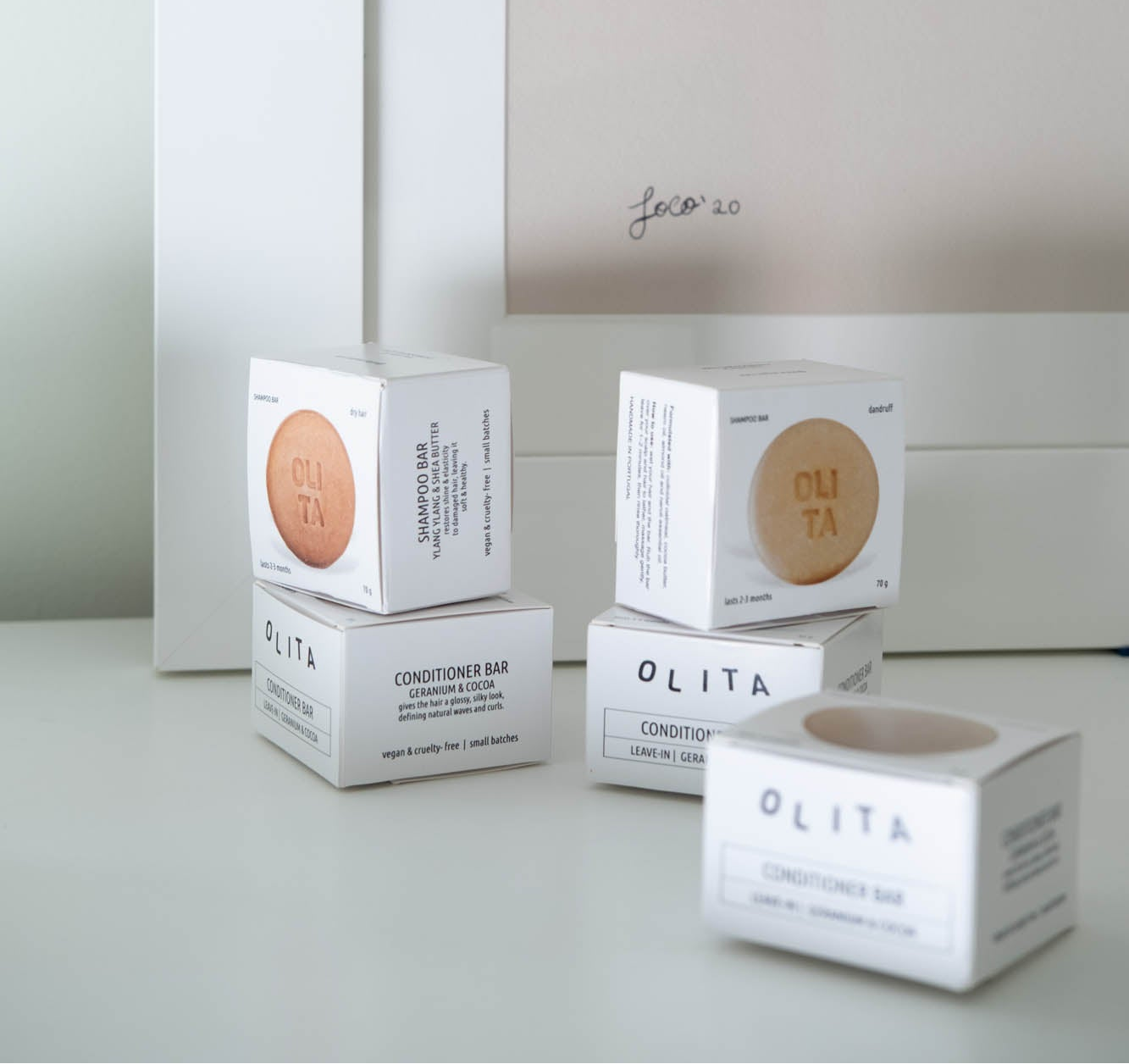 Several OLITA Conditioner Bar boxes are arranged on a light gray surface against a pale beige wall. The white packaging shows the circular amber-brown conditioner bar through cutouts on the front faces.