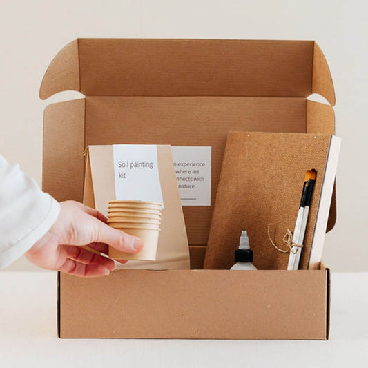 An open kraft cardboard box containing a self-painting kit, including paper cups, a notebook, a marker, and a small bottle, with a hand reaching in to grab one of the cups against a clean white background.