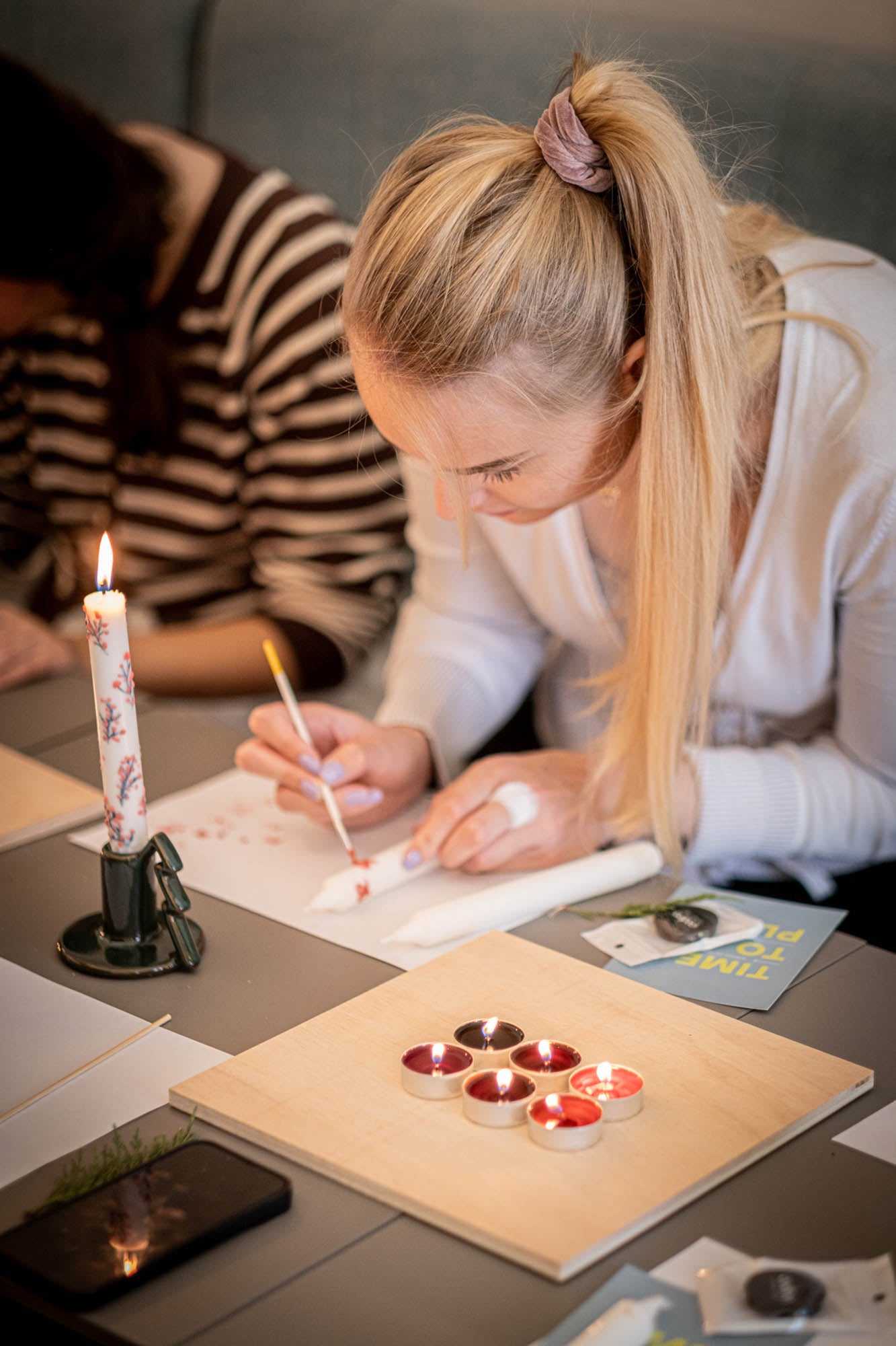 Woman painting with watercolors at a desk with a candle and paint supplies.