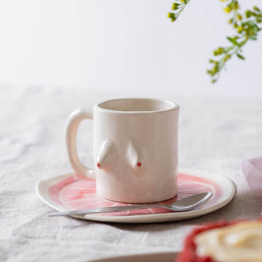A beige ceramic mug with a simplistic design resembling a breast, placed on a matching beige and pink plate on a table with a floral ornament in the background.