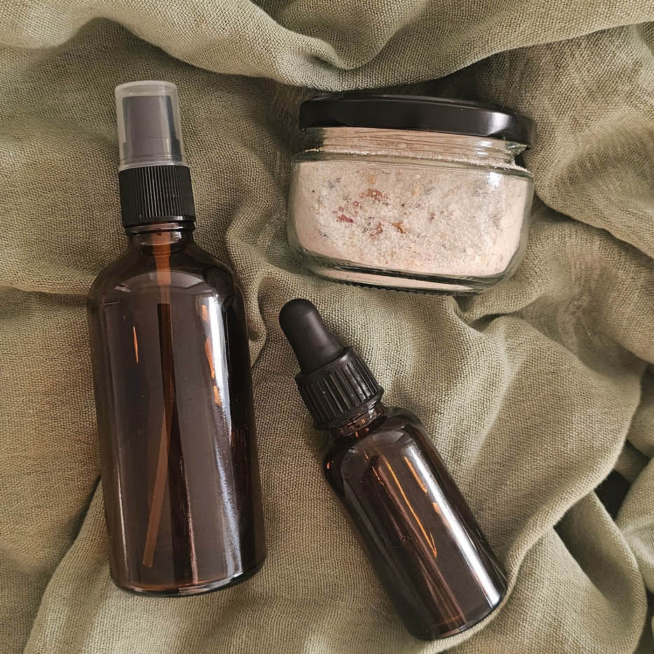 Three glass bottles with different accessories on a textured green fabric background: exfoliant, cleanser and serum.