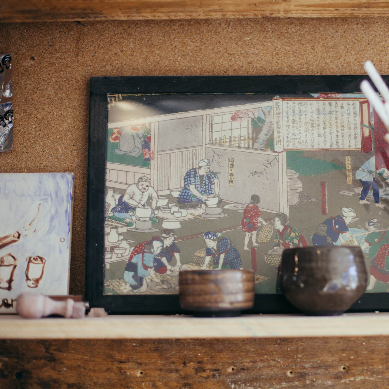 Framed artwork on a wooden shelf with various items including a mug and brushes.