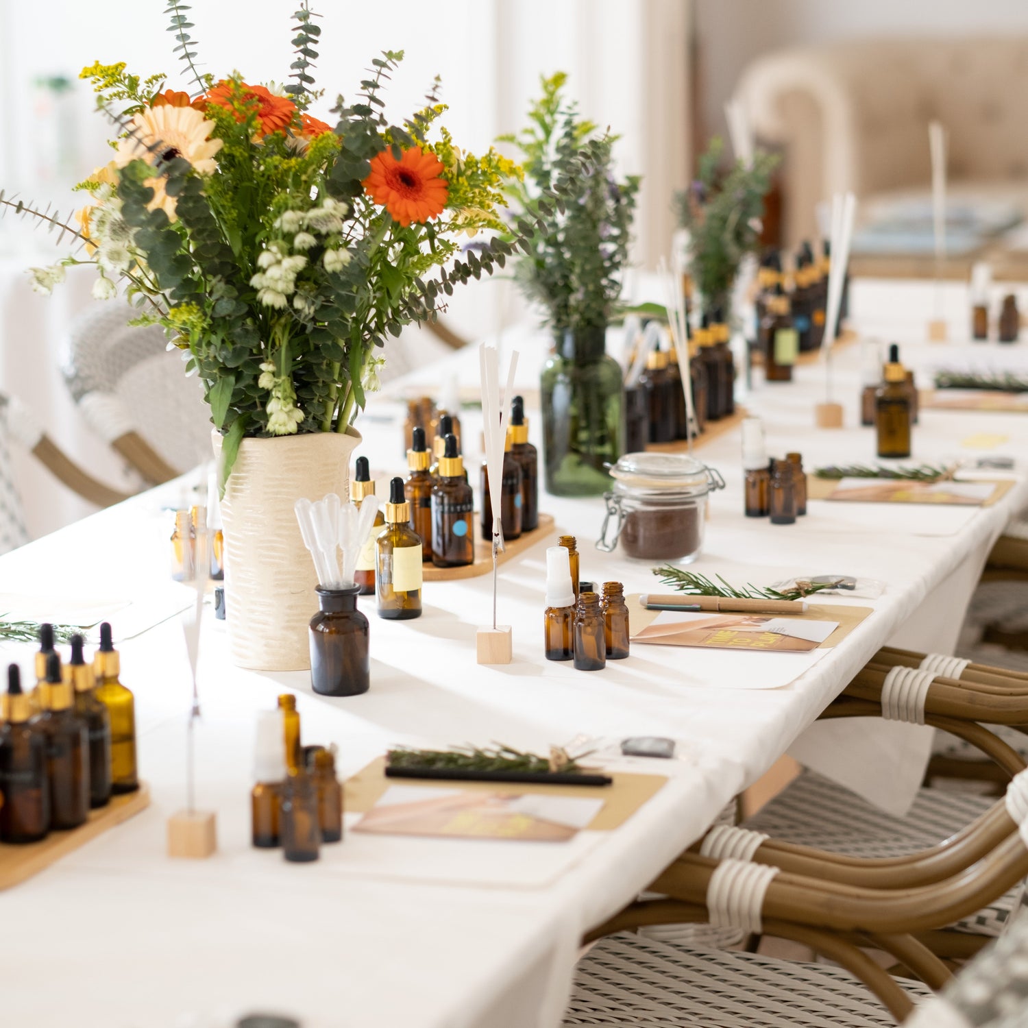 A styled event table setting featuring numerous amber glass dropper bottles, floral centerpieces with orange flowers and green foliage, woven chairs, and a white tablecloth in a bright indoor setting.