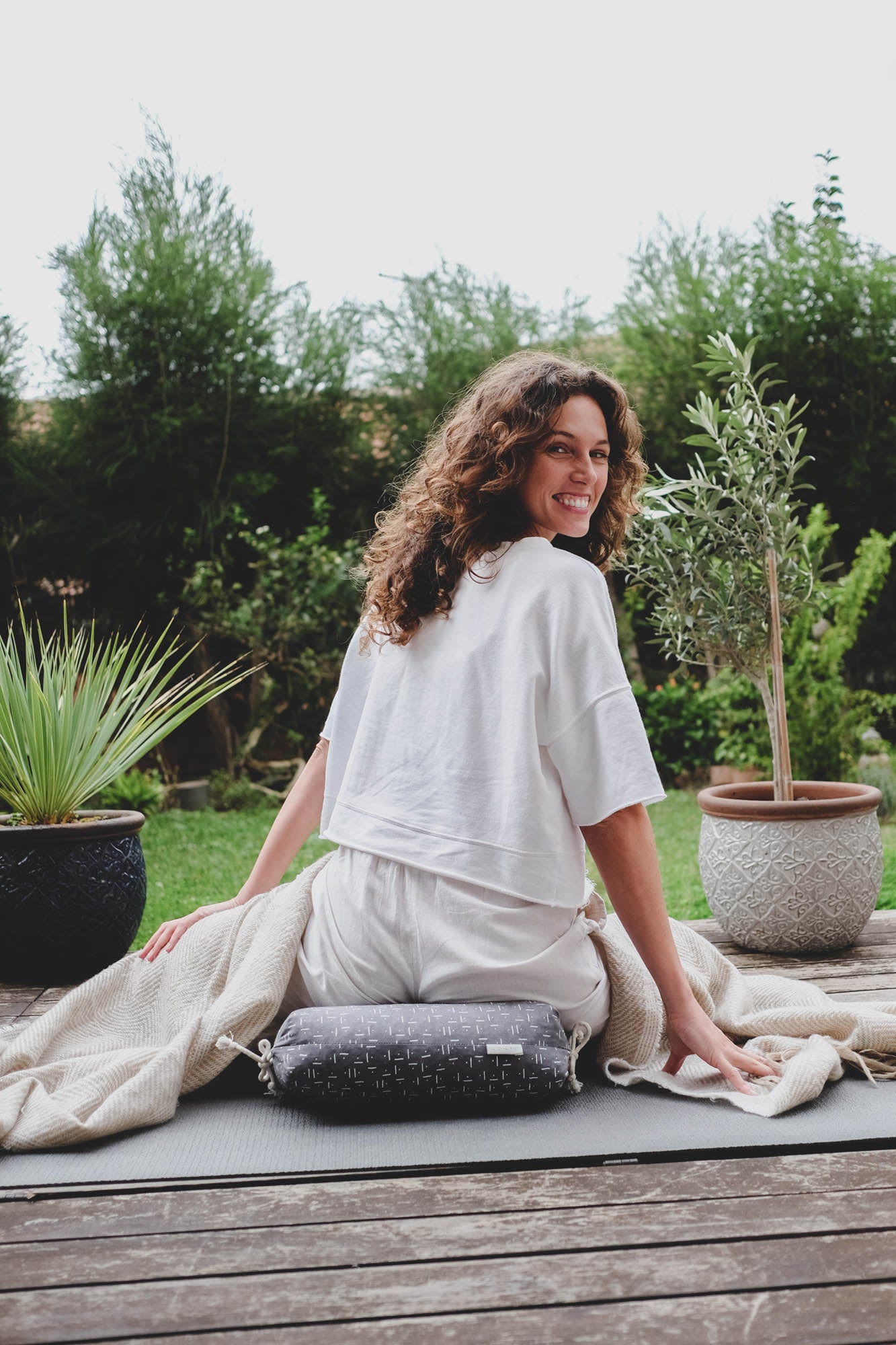 Woman sitting on zafu in a wooden deck with plants and a blanket