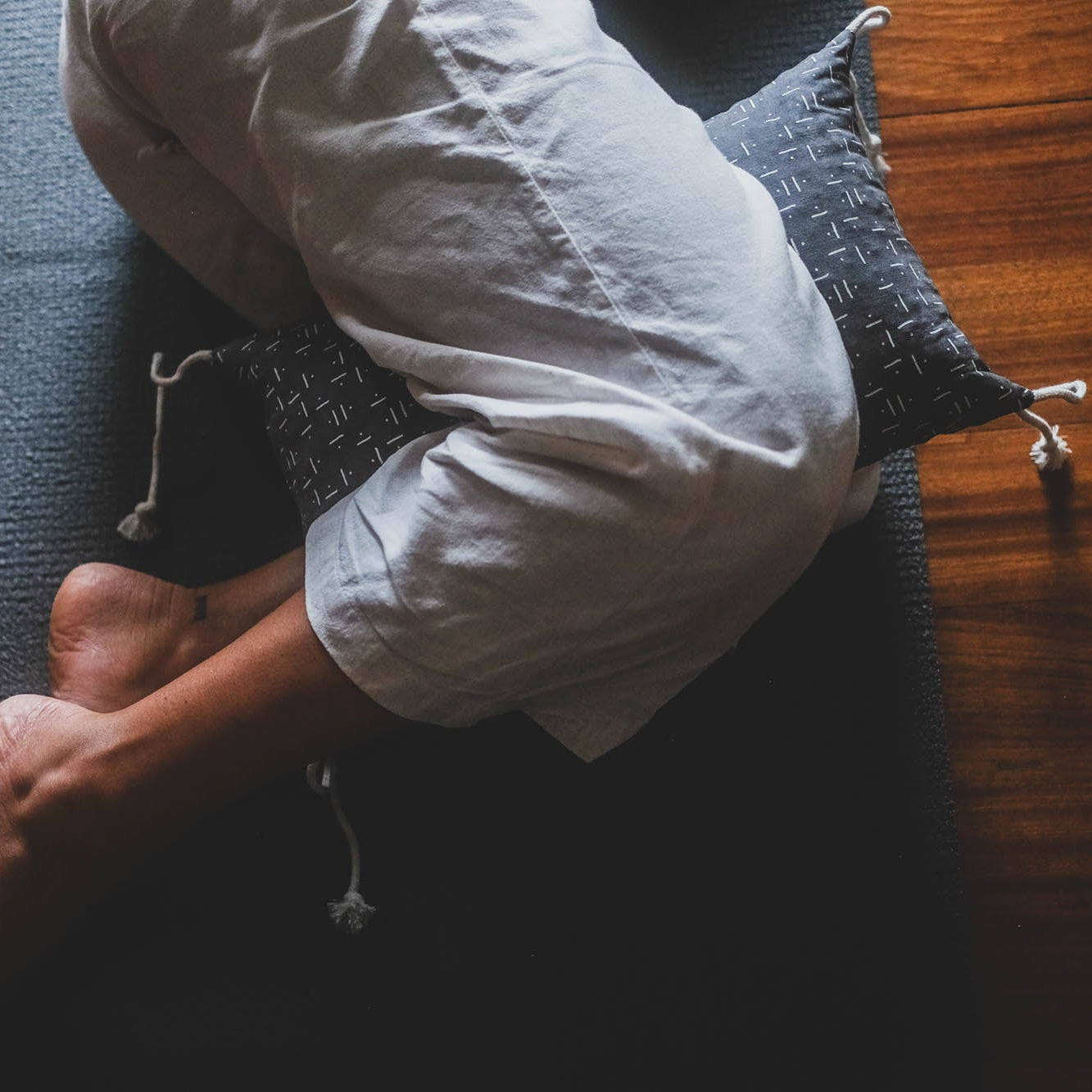 Person sitting on a yoga mat with a pillow under their head on a wooden floor.