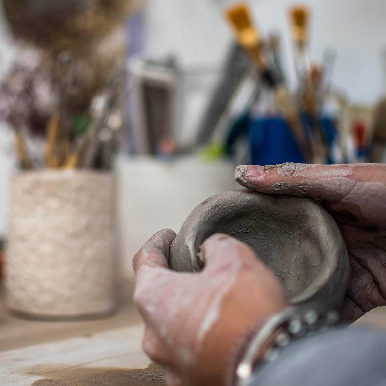 Ceramic Workshop in Lisbon, Portugal. Close-up of a person's hands shaping a small gray clay bowl in a pottery studio, with blurred brushes and tools in the background.