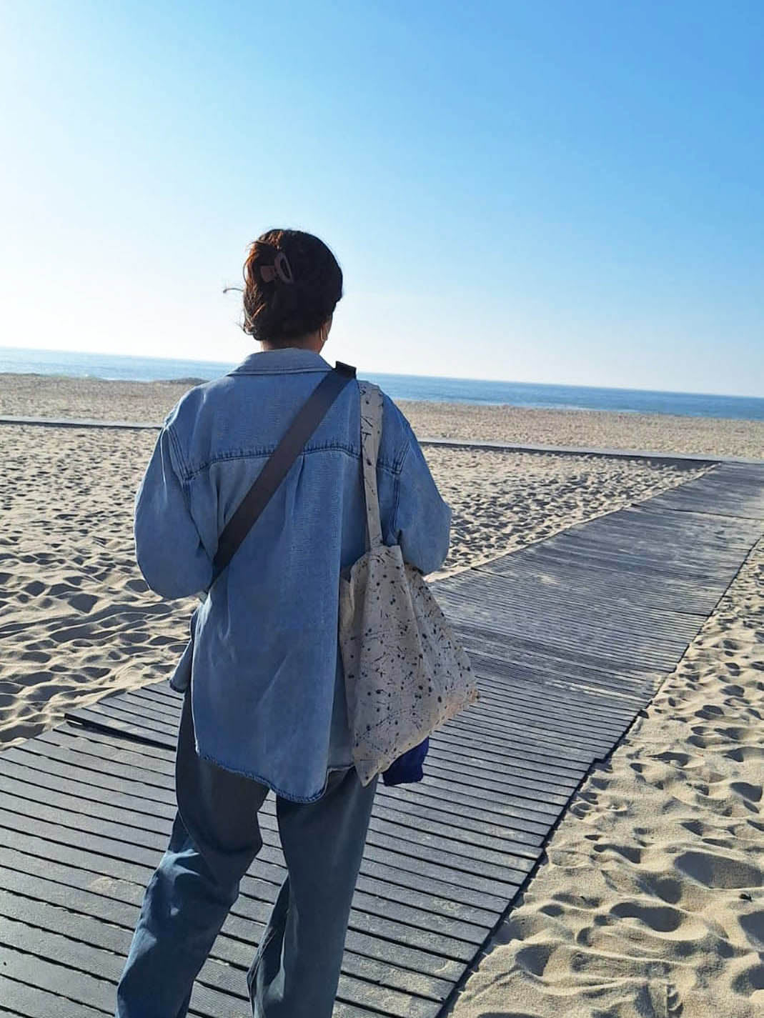 A person viewed from behind standing on a wooden boardwalk at the beach, wearing a light blue denim shirt and dark pants, with a patterned eco printed tote bag slung over the shoulder.