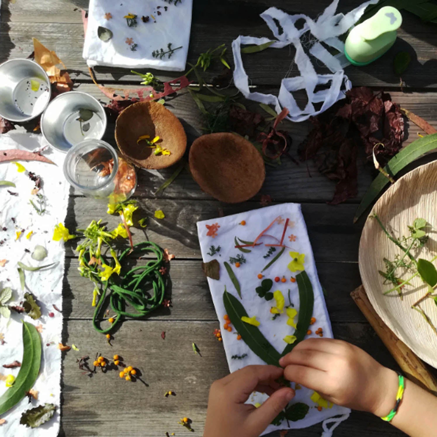 A top-down view of a handmade flower pressing station on a rustic wooden table. Hands are visible arranging pressed green leaves and small yellow flowers onto white paper sheets, surrounded by tools like wooden bowls, metal cups, and dried botanicals.