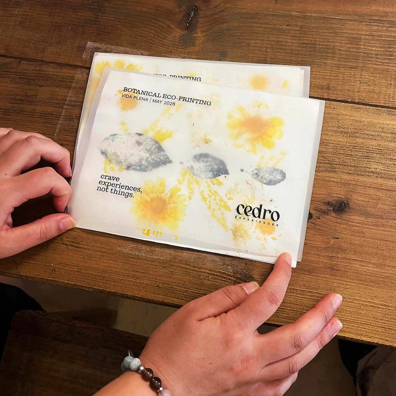 A person's hands holding botanical  print postcards on a rustic wooden table. The print features yellow flowers and gray stone-like textures with the text 'cedro eco artistry' and 'botanical eco-printing'.