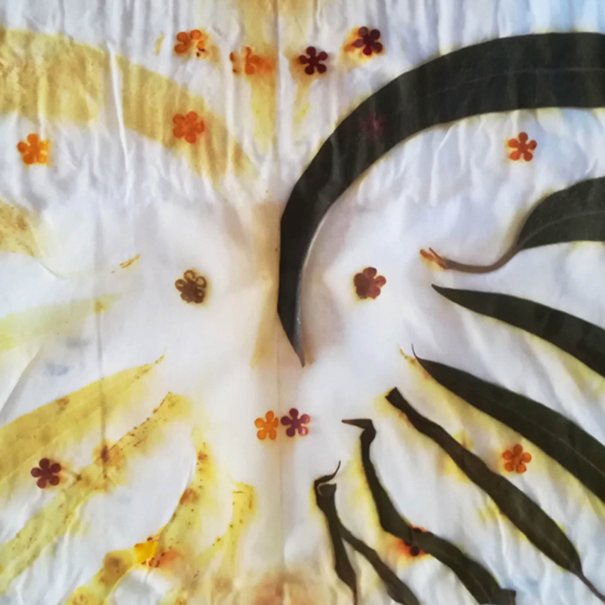 A close-up of a white fabric with yellow tie-dye or batik patterns, decorated with pressed botanical elements including dark green leaves and small orange-brown flowers.
