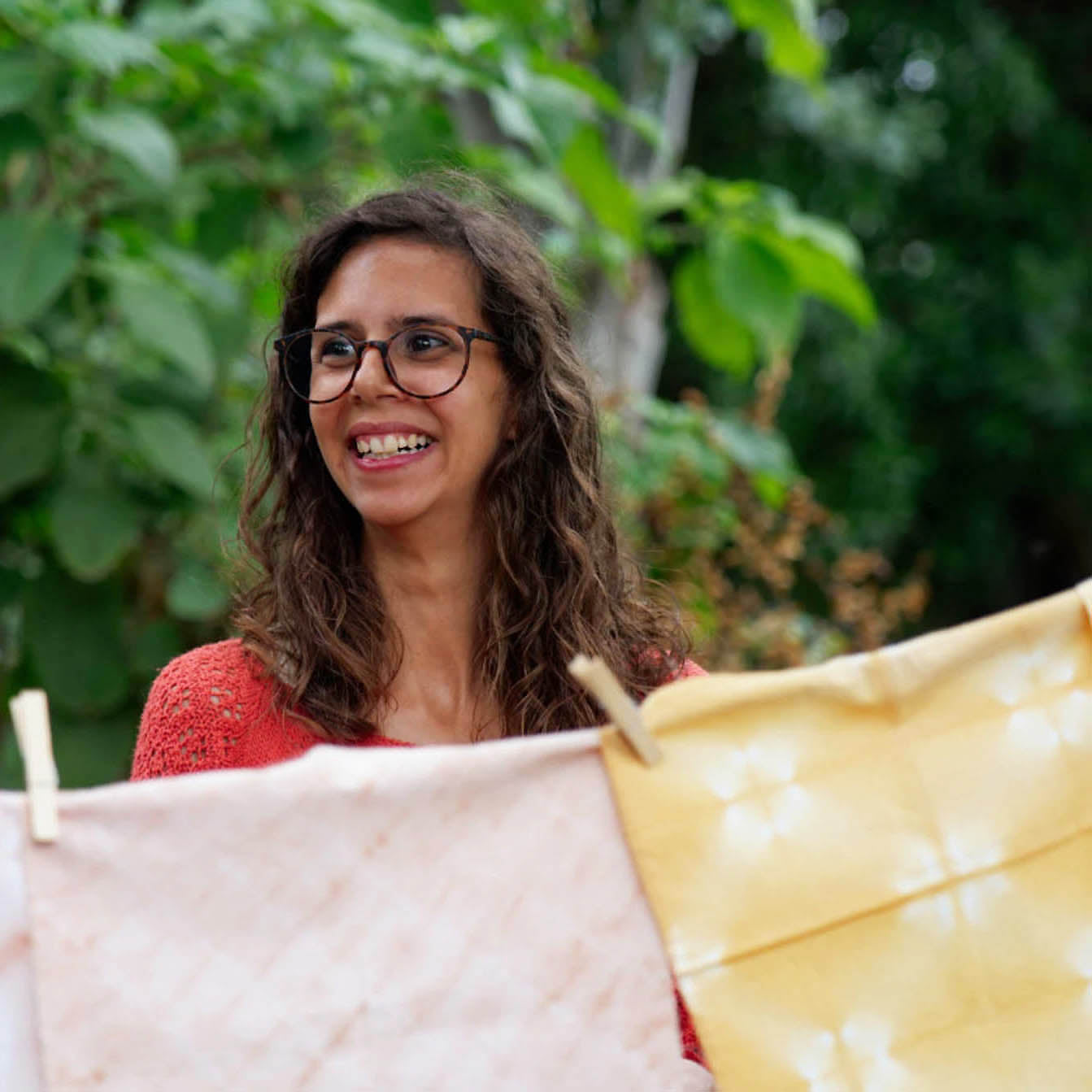 A woman with glasses and brown hair smiles while holding up two fabric cloths, one light pink and one yellow, clipped together like a clothesline, with a green garden background.