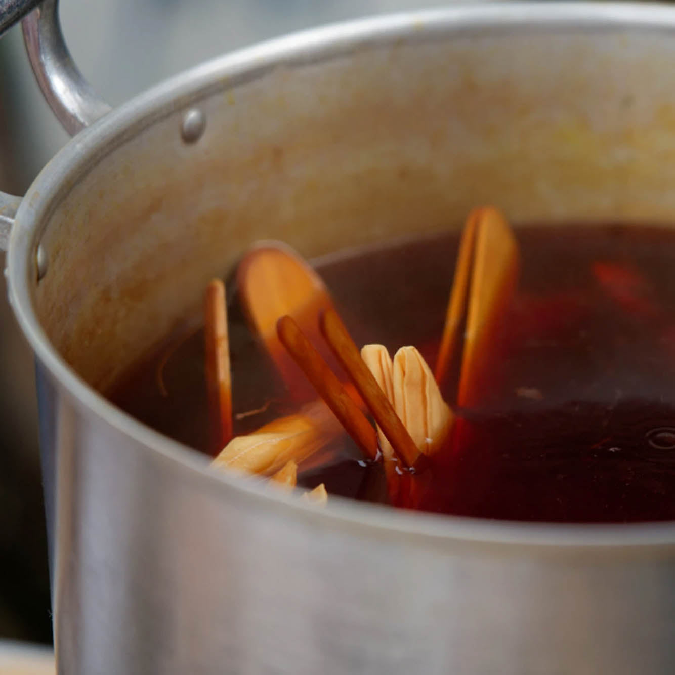 A close-up of a metal pot containing dark reddish-brown liquid with several light brown wooden sticks or strips partially submerged.