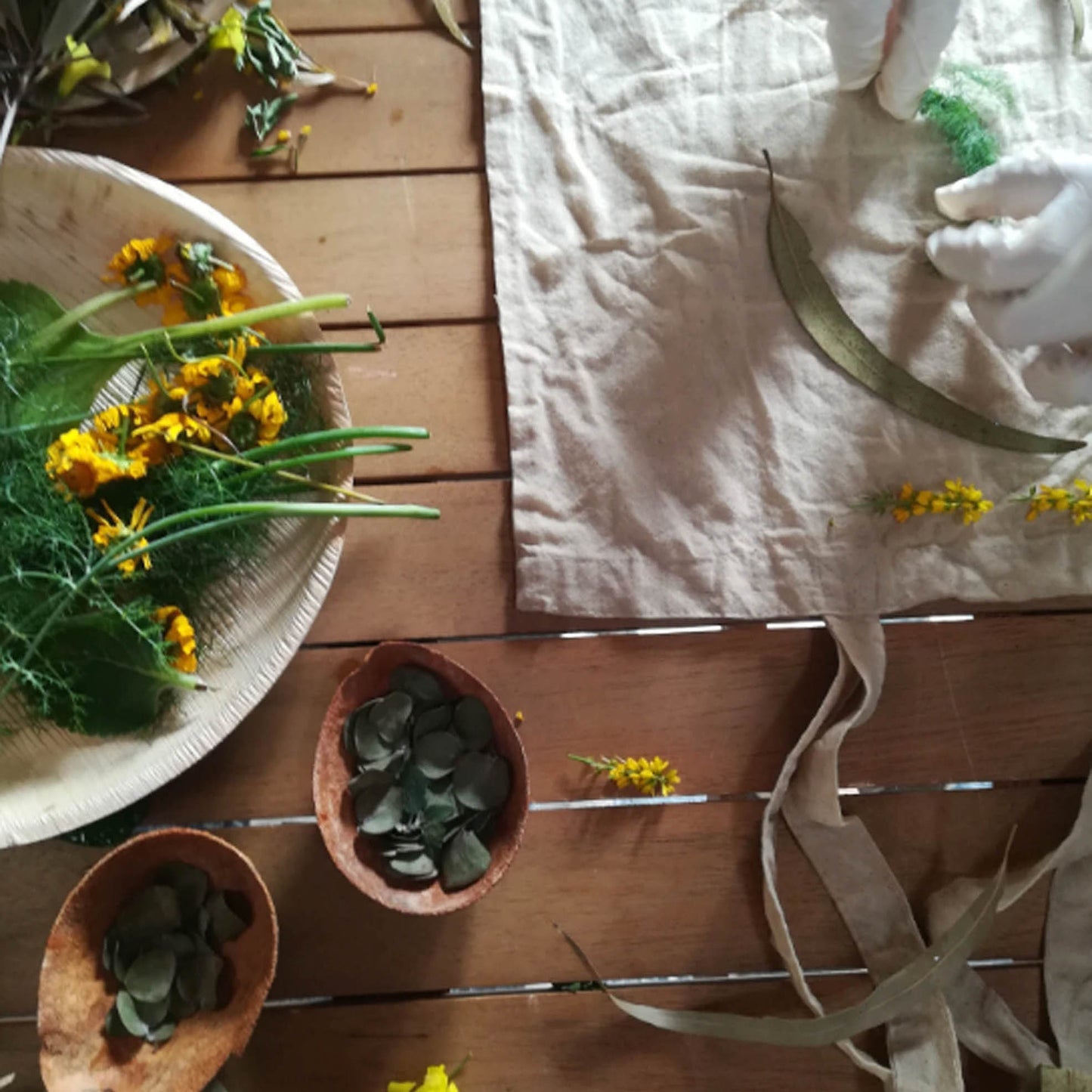 A styled flat-lay of a beige linen tote bag on a wooden table, surrounded by natural elements including yellow wildflowers, green leaves, and small bowls containing dark green stones.