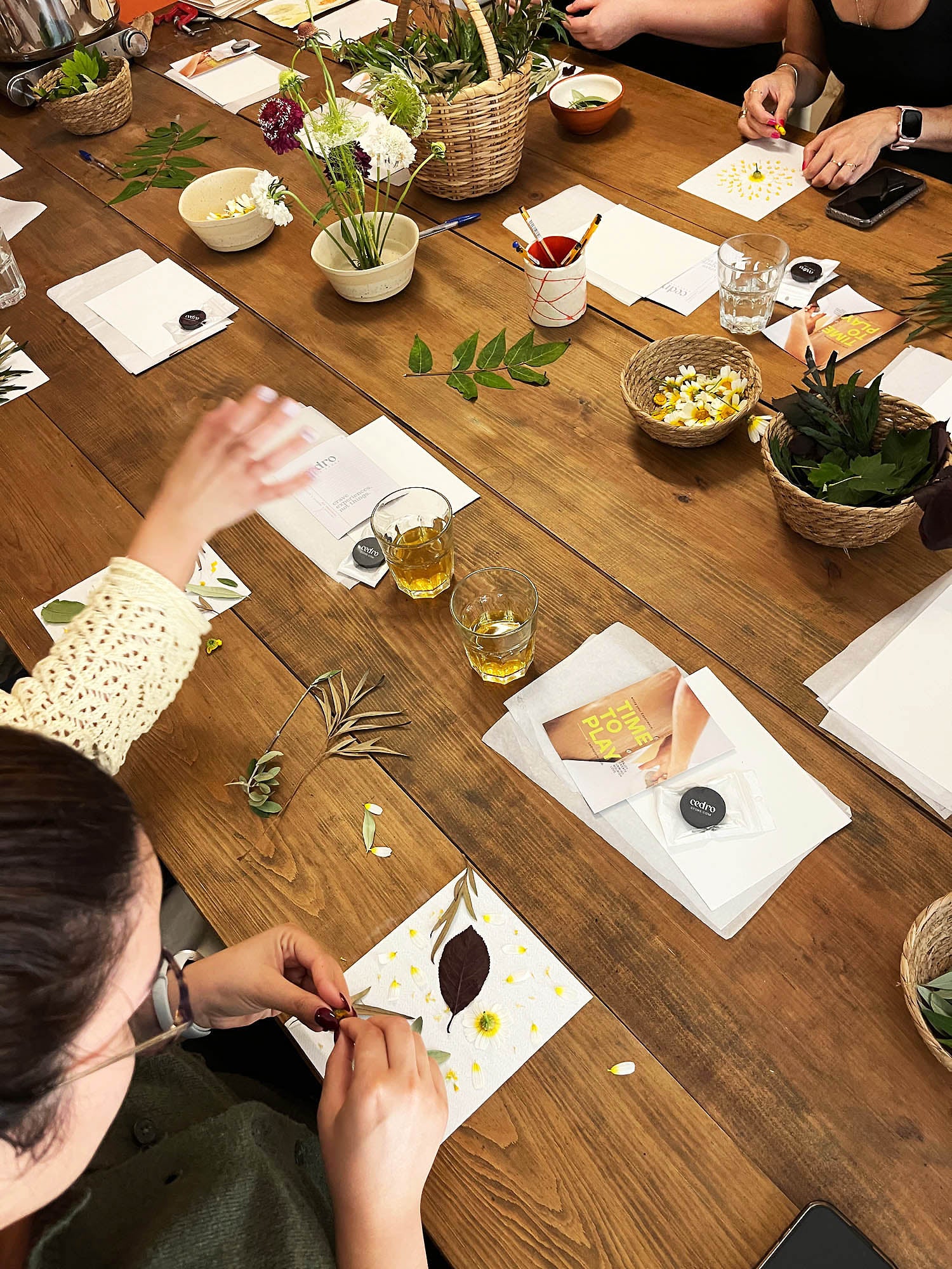 A group of people seated around a rustic wooden table, engaged in a botanical art or craft workshop. Papers with pressed flowers, small woven baskets, and floral arrangements are spread across the table.