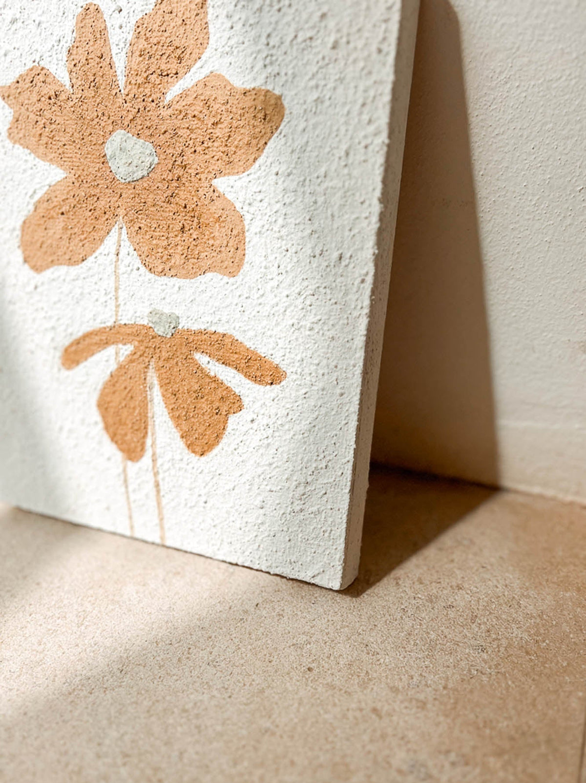 A close-up of a textured white wall art panel leaning against a beige wall on a beige floor, featuring a large and a small hand-painted style tan flower with a small white center.