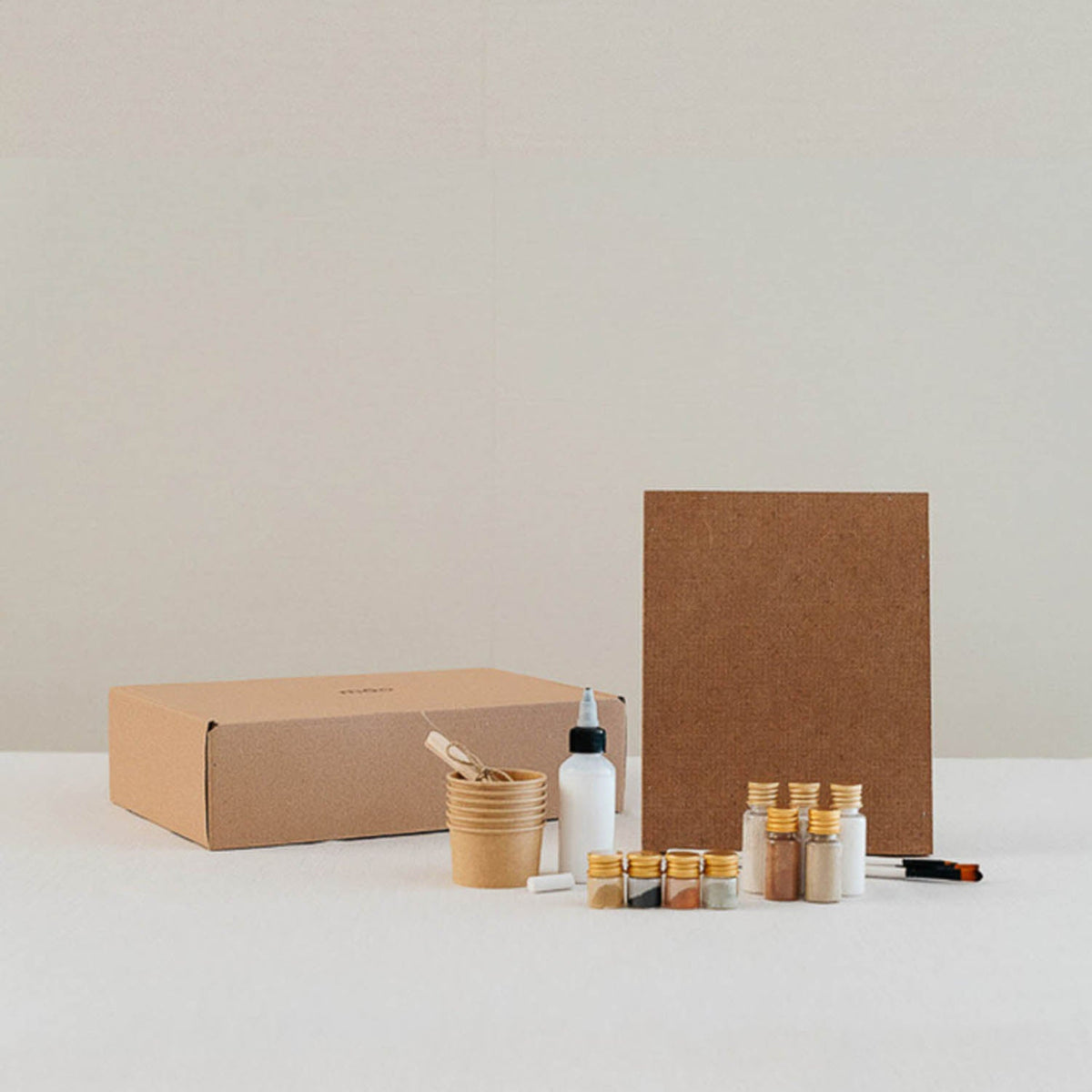 A minimalist craft or art kit arranged on a white surface against a light beige wall, featuring a brown kraft box, a brown paper board, small glass vials with gold caps, a white bottle, a cup with tools, and a small black marker.