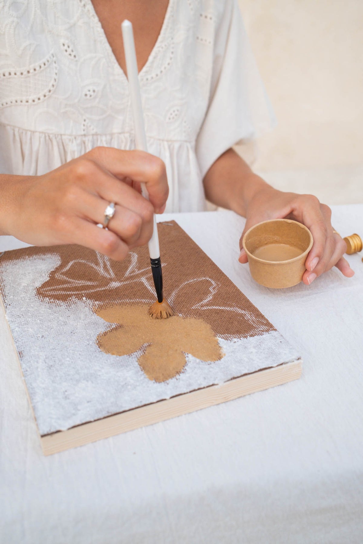 A person wearing a white embroidered blouse is painting a floral design on a canvas. Their right hand holds a fine-tip paintbrush applying brown paint to a flower, while their left hand holds a small cup of water.