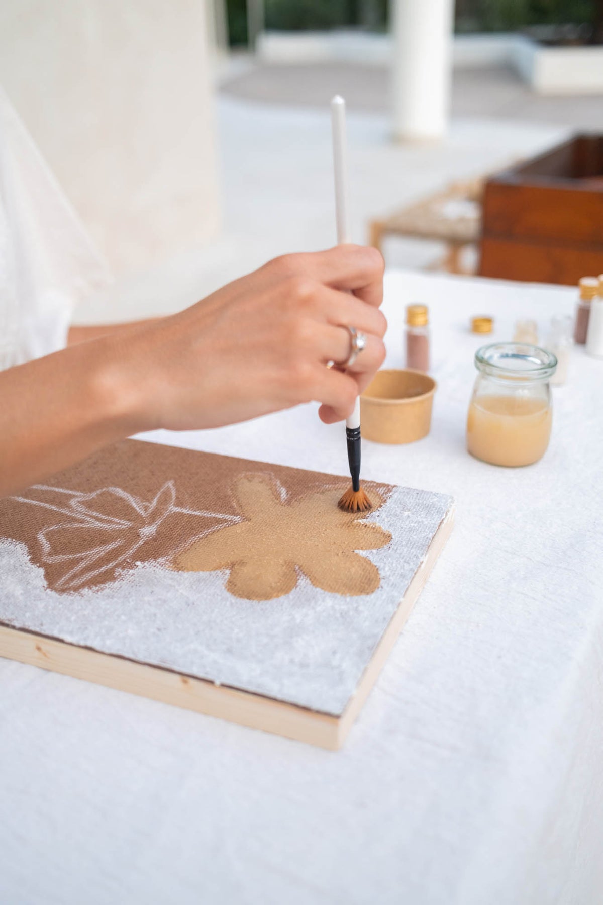 A close-up of a hand holding a paintbrush and painting a tan leaf design onto a textured canvas or board, set on a white tablecloth with small jars of paint in the background.