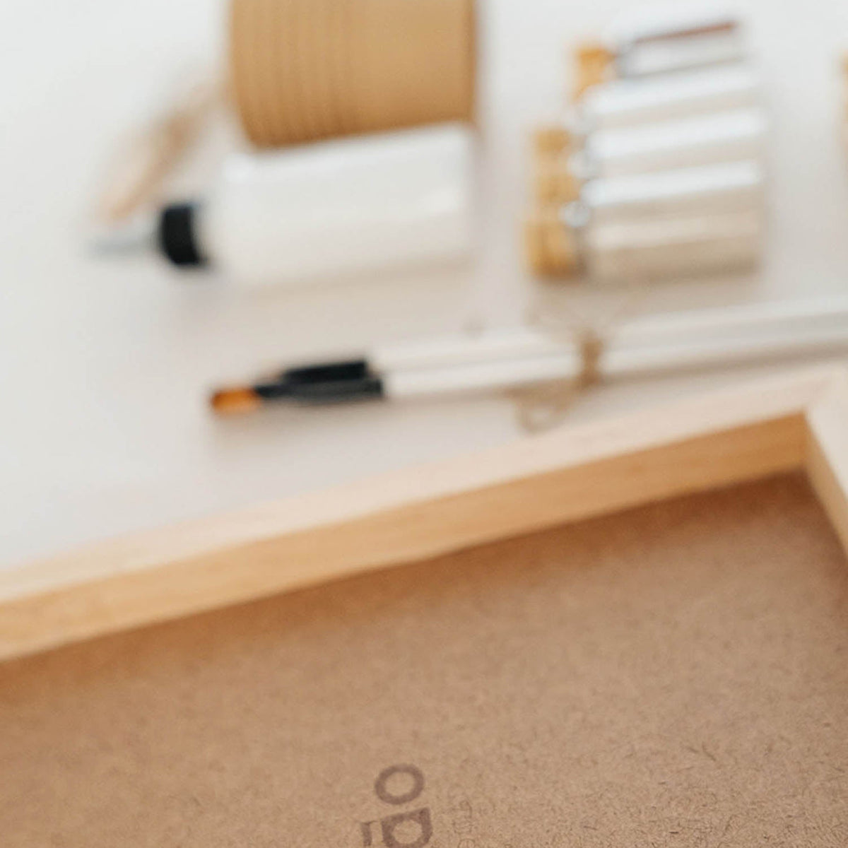 A close-up of a light wood tray or frame with a brown kraft-paper insert printed with the word 'mão' in lowercase. A white pen and small cosmetic-like items are blurred in the background on a white surface.