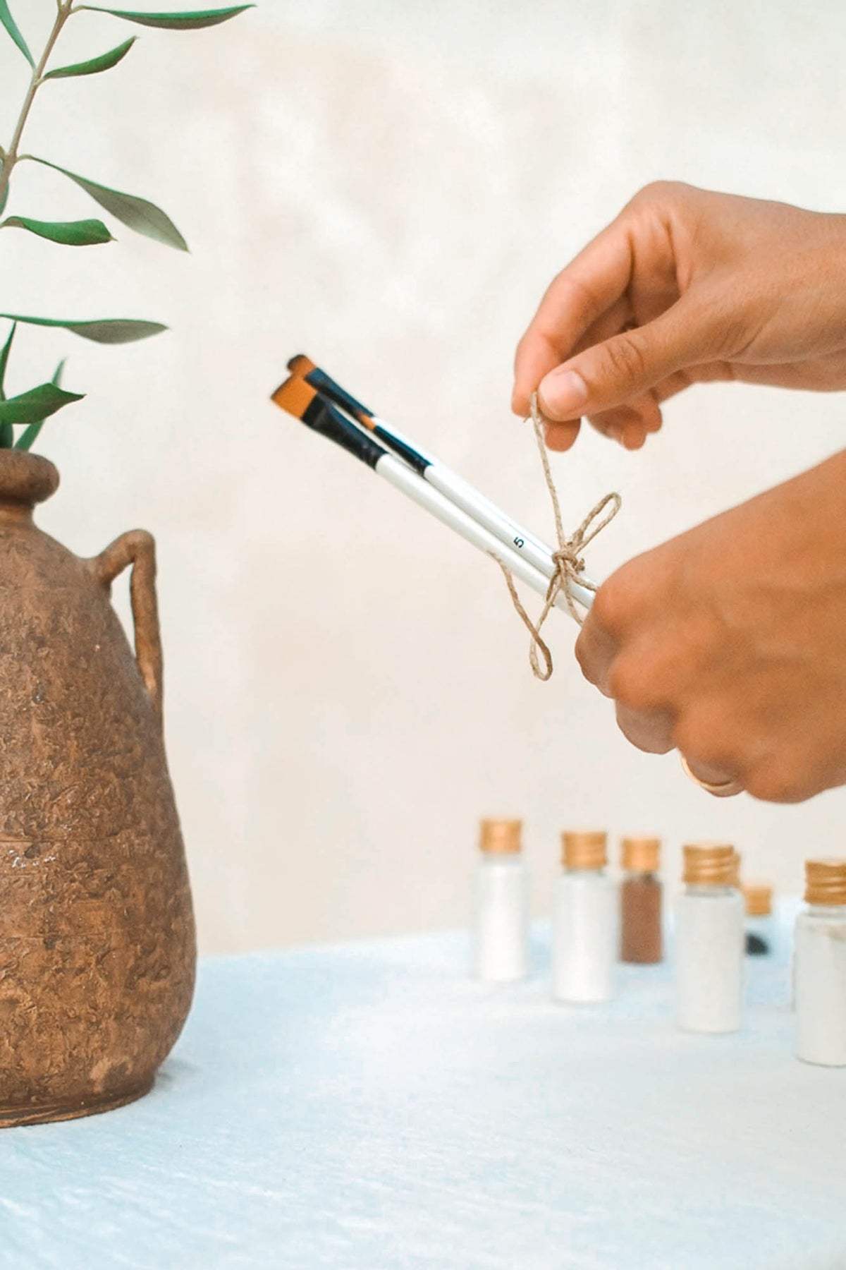 A close-up of a person's hands holding two white paintbrushes tied together with twine, with a terracotta vase and small glass vials in the background.
