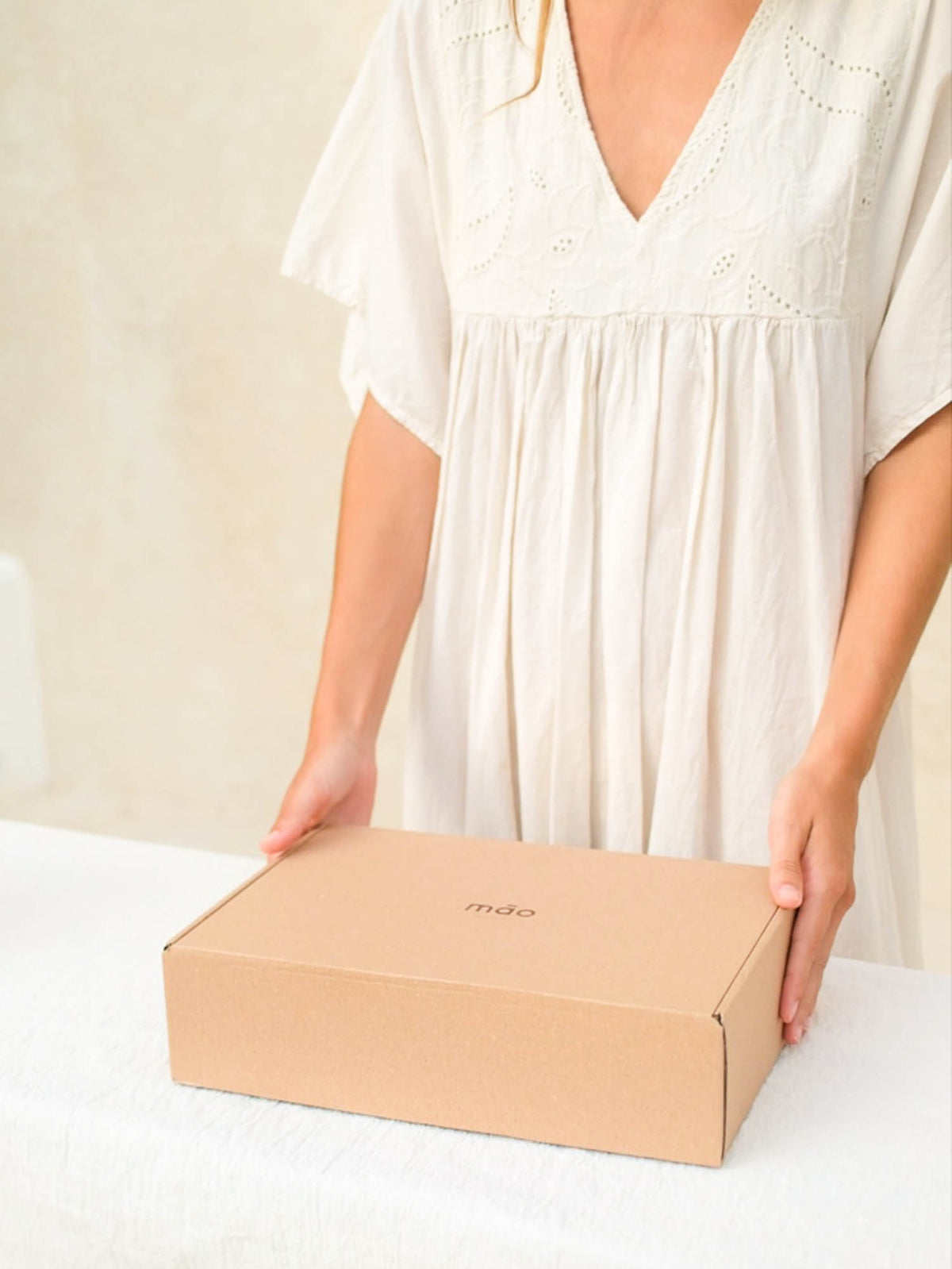 A person in a white eyelet dress holds a plain brown cardboard box on a white surface against a light beige wall.