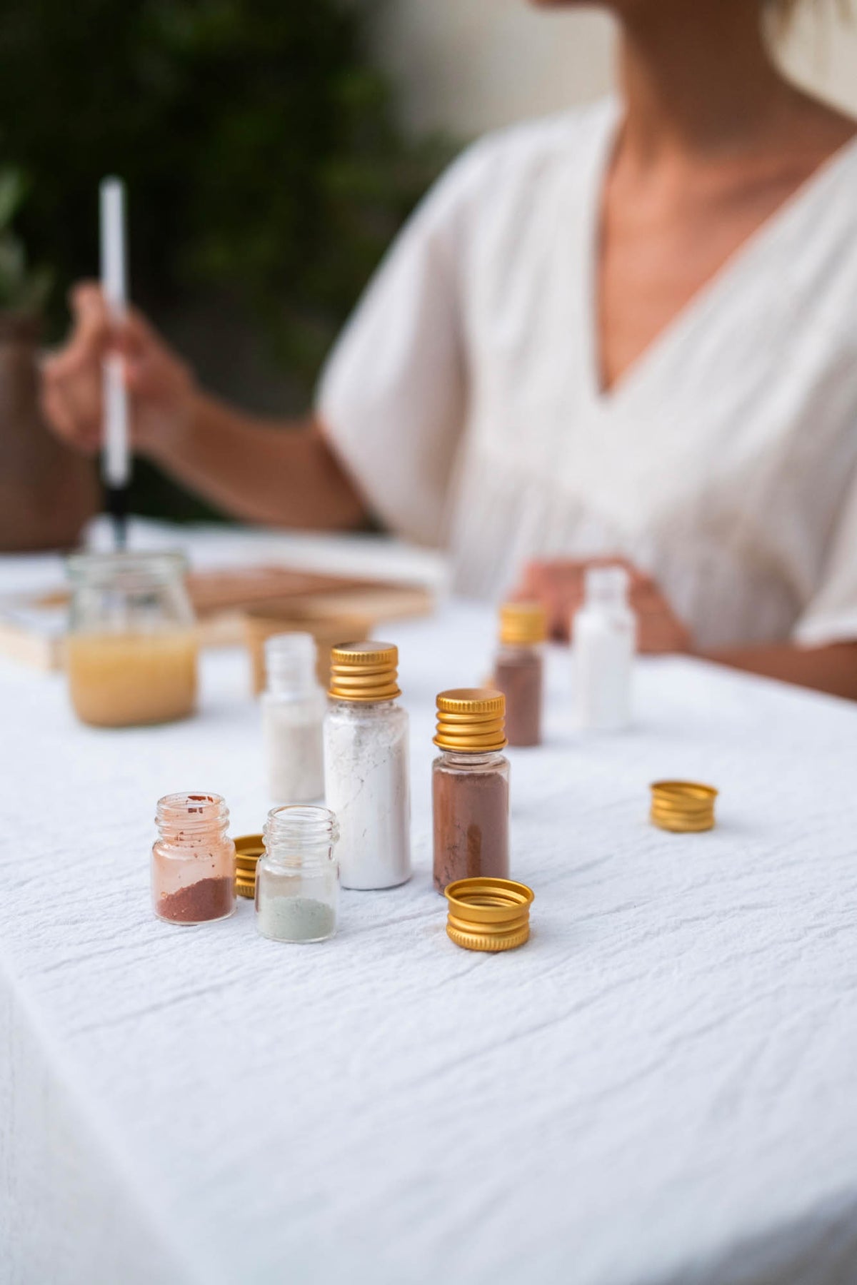 Small glass jars with gold caps containing white, brown, and pink powders arranged on a white tablecloth, with a person in a white top holding a brush in the softly blurred background.