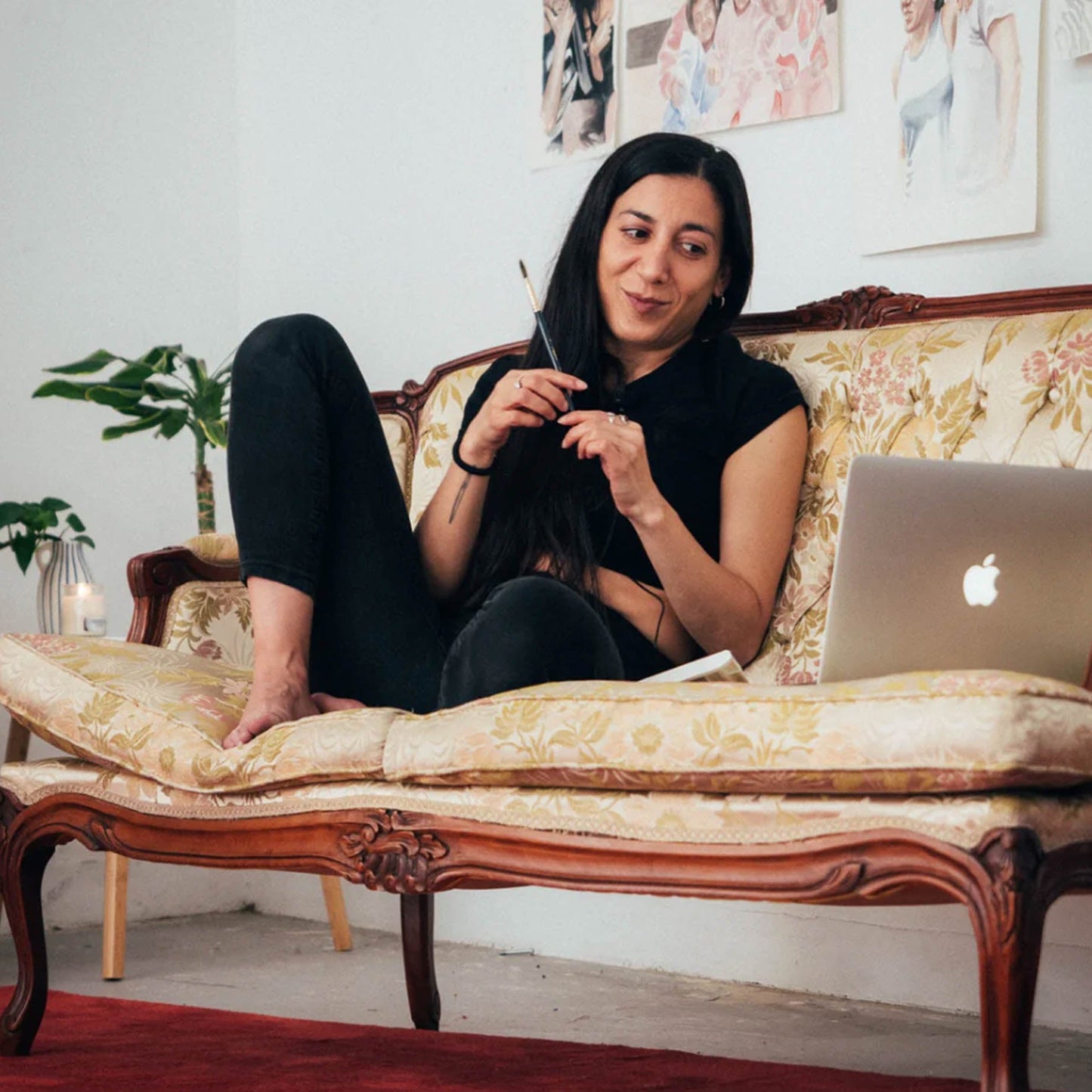 Woman sitting on a floral-patterned couch with a laptop, surrounded by decorative elements.