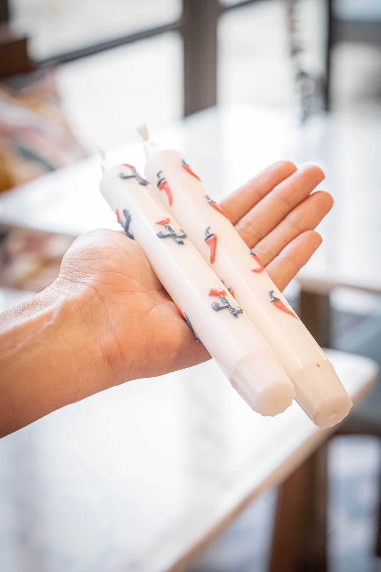 Hand holding two white candles with decorative elements against a blurred indoor background