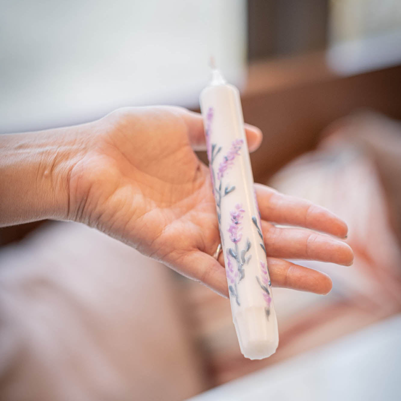 Hand holding a white candle with floral  - lavander- design against a blurred background