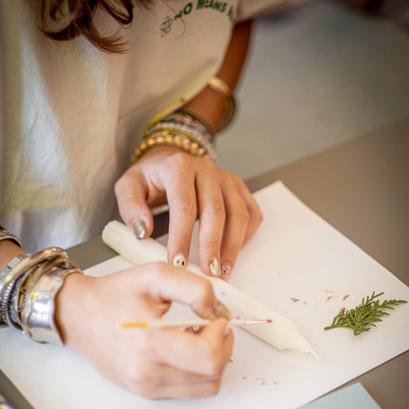 Person's hands with bracelets and nail polish working on a craft project with paper and greenery.