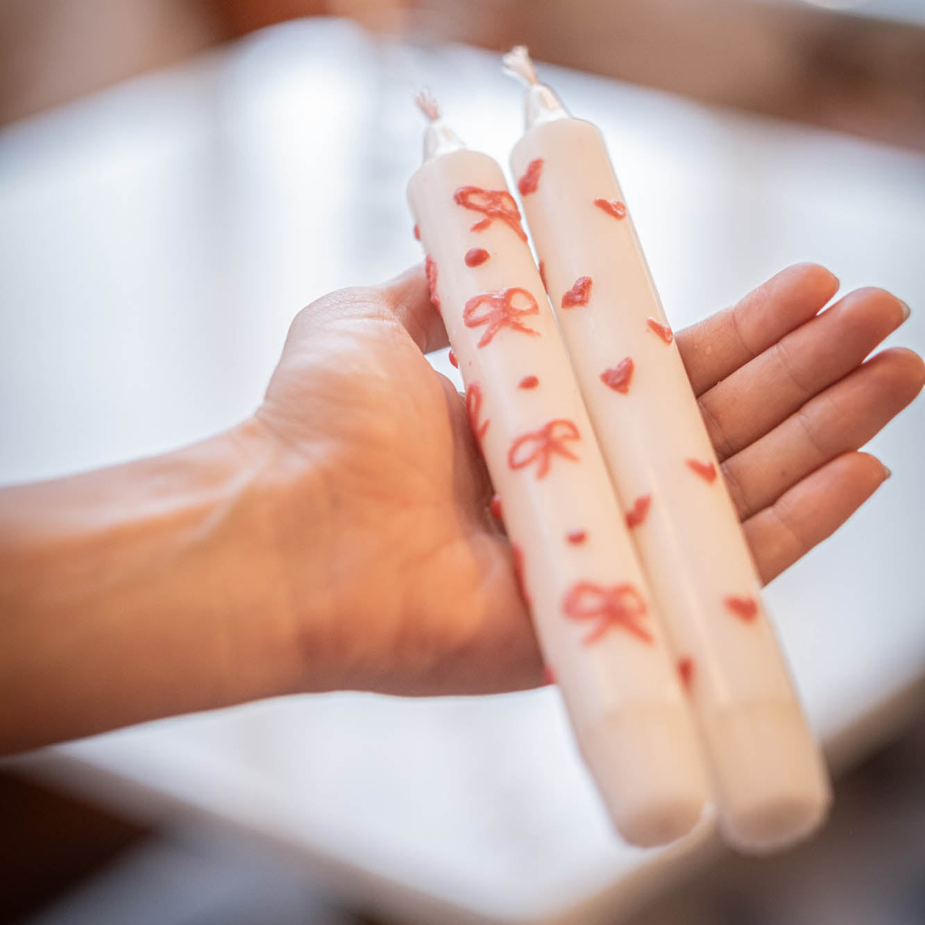 Two candles with heart and bow designs held in a hand against a blurred background
