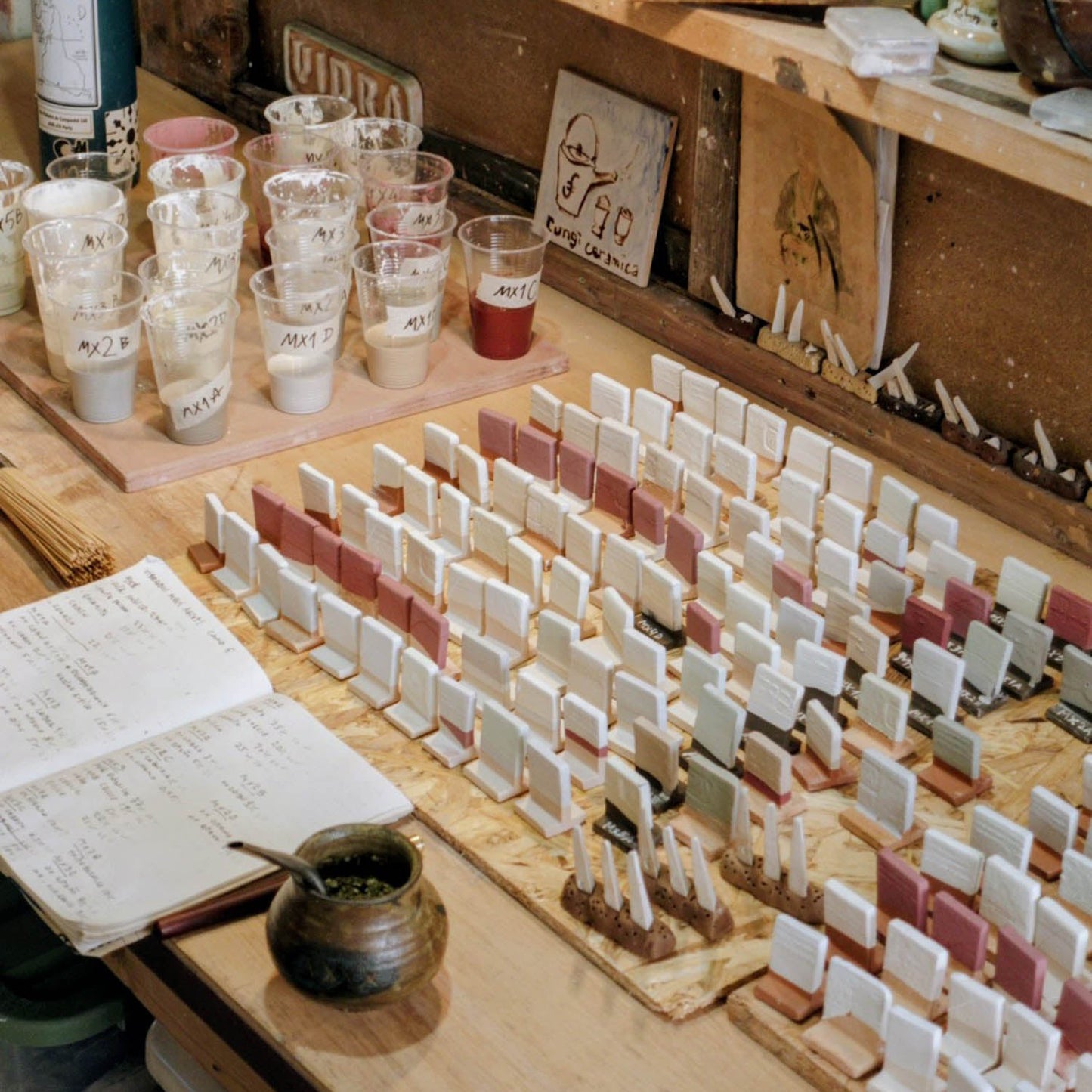 Workshop table with ceramic glazes tests, tools, and supplies on a wooden shelf.