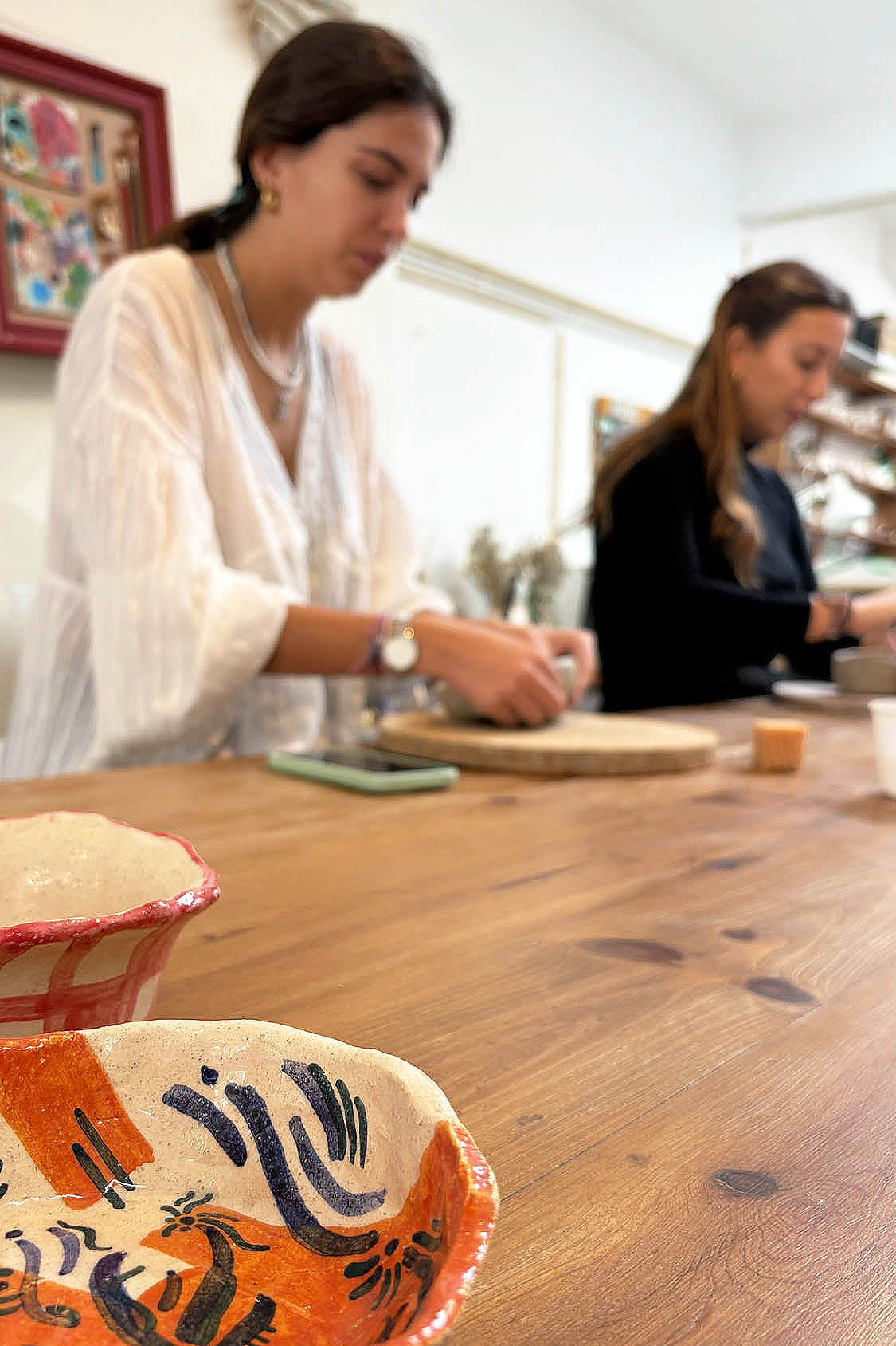 Ceramic Workshop in Lisbon, Portugal. A woman in a white blouse stands at a wooden table in a bright indoor setting, working with clay or pottery. Another person is blurred in the background.