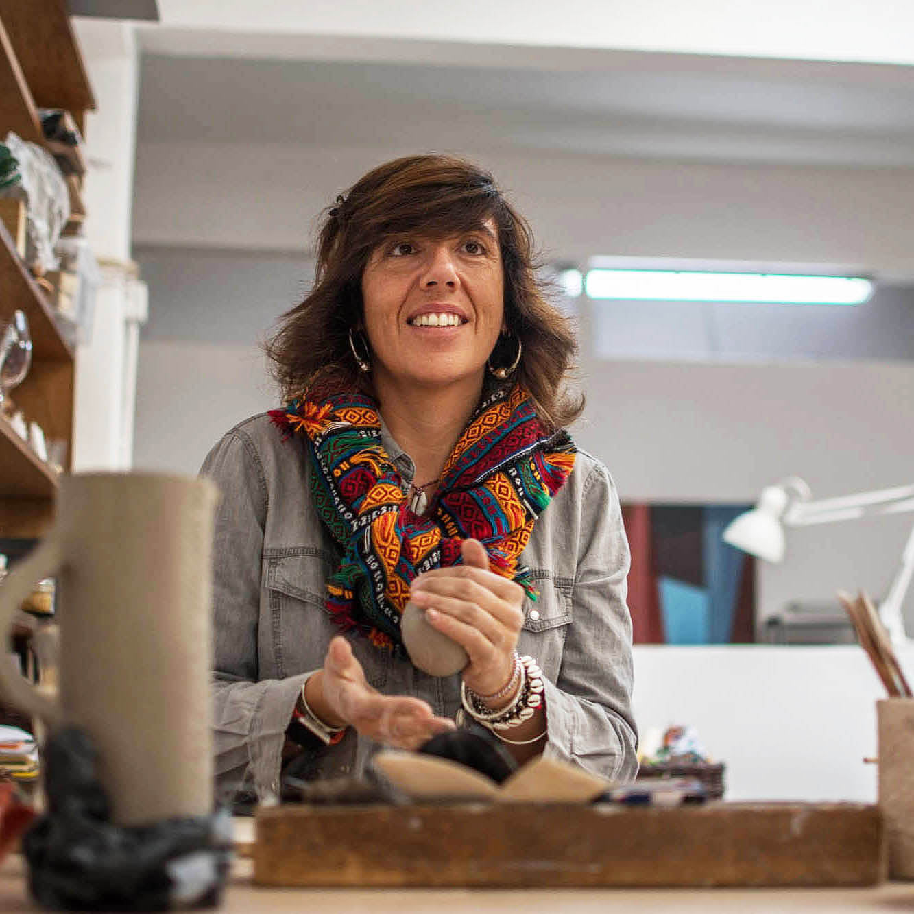 Ceramic Workshop in Lisbon, Portugal. THE HOST: a smiling woman with dark hair stands behind a wooden table in a bright studio or workshop, wearing a gray denim shirt and a colorful patterned scarf. 