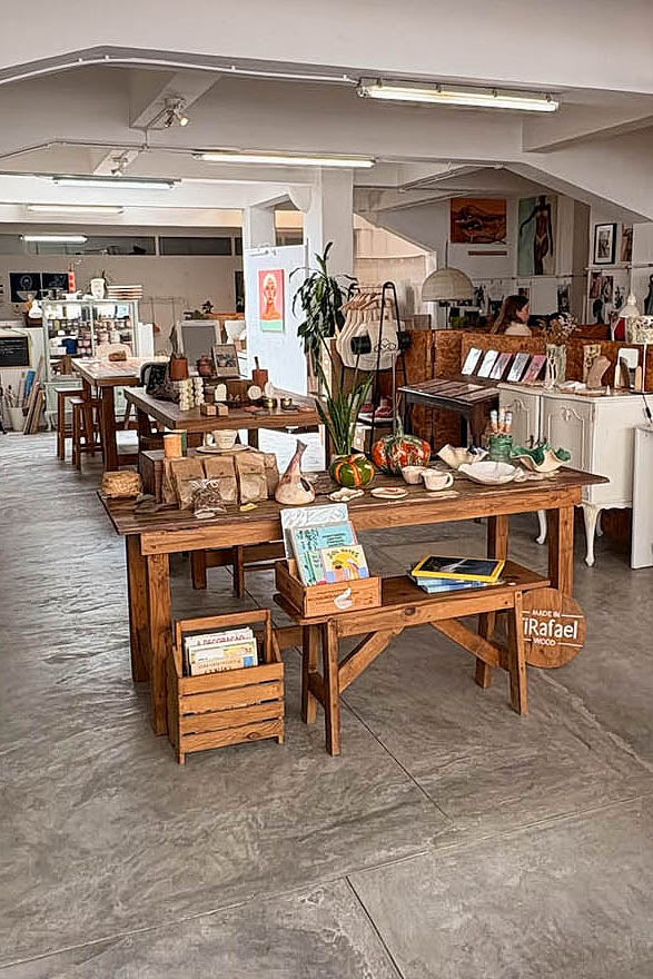 Ceramic Workshop in Lisbon, Portugal. THE ATELIER: a rustic wooden display table in a vintage or antique shop, styled with books, small decorative items, and potted plants on a gray concrete floor.