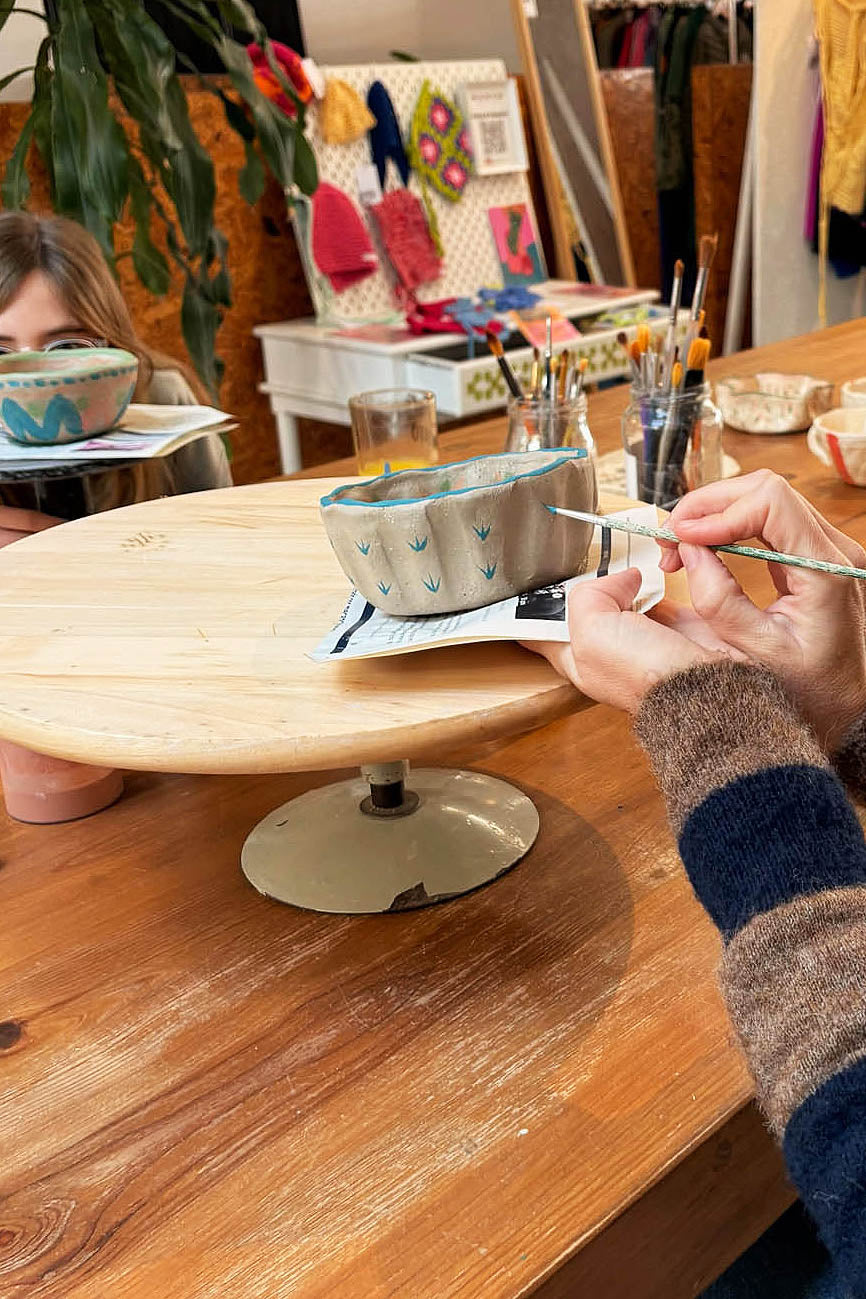 Ceramic Workshop in Lisbon, Portugal. A person painting a small ceramic bowl on a wooden spinning wheel in a pottery studio. The bowl has a light gray base with a blue rim and small green leaf patterns.