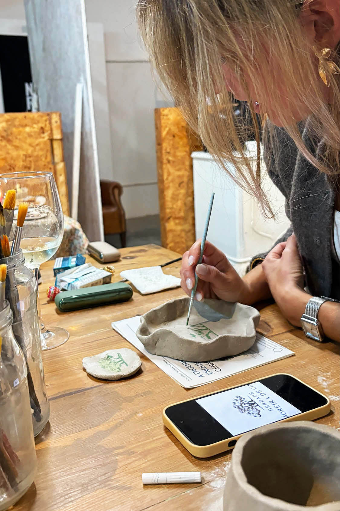 Ceramic Workshop in Lisbon, Portugal. A woman with blonde hair is shown from the side, carefully painting a small handmade clay dish on a wooden table. The table is cluttered with pottery tools, brushes, a wine glass, a smartphone, and other clay pieces, indicating a pottery workshop or studio environment.