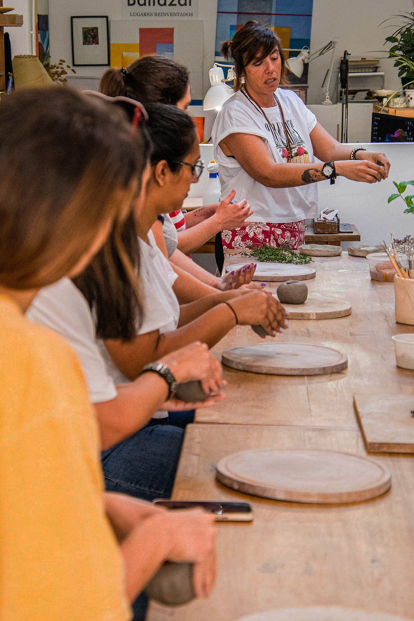 Ceramic Workshop in Lisbon, Portugal. A pottery class in progress with a woman demonstrating while several students sit at a long wooden table, each holding a clay wheel.