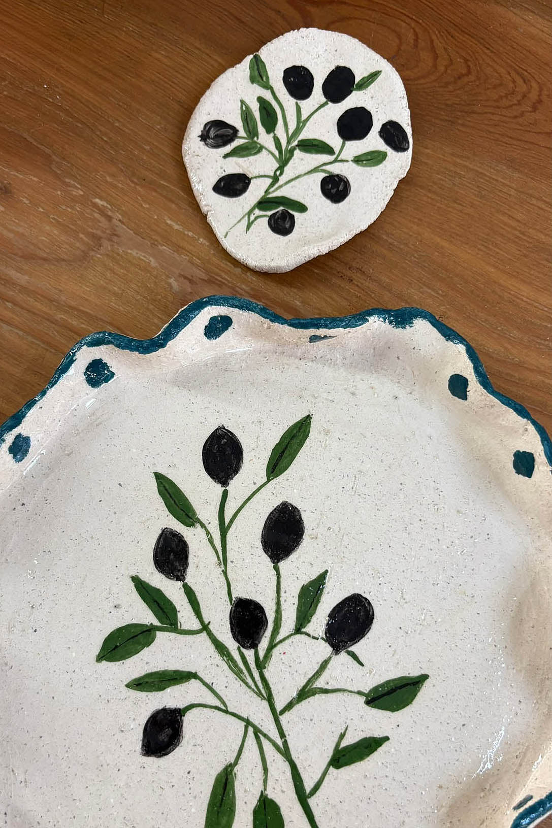 Ceramic Workshop in Lisbon, Portugal. A handmade ceramic olive branch plate on a wooden table, featuring a white speckled glaze, a scalloped blue rim, and a painted green olive branch motif with black olives. A matching small round coaster or tile is placed above the plate.