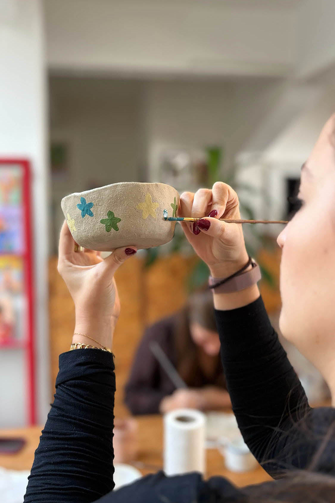 Ceramic Workshop in Lisbon, Portugal. A person holding a small beige ceramic cup with painted green and blue flowers, using a paintbrush to apply paint, in a softly blurred indoor setting.