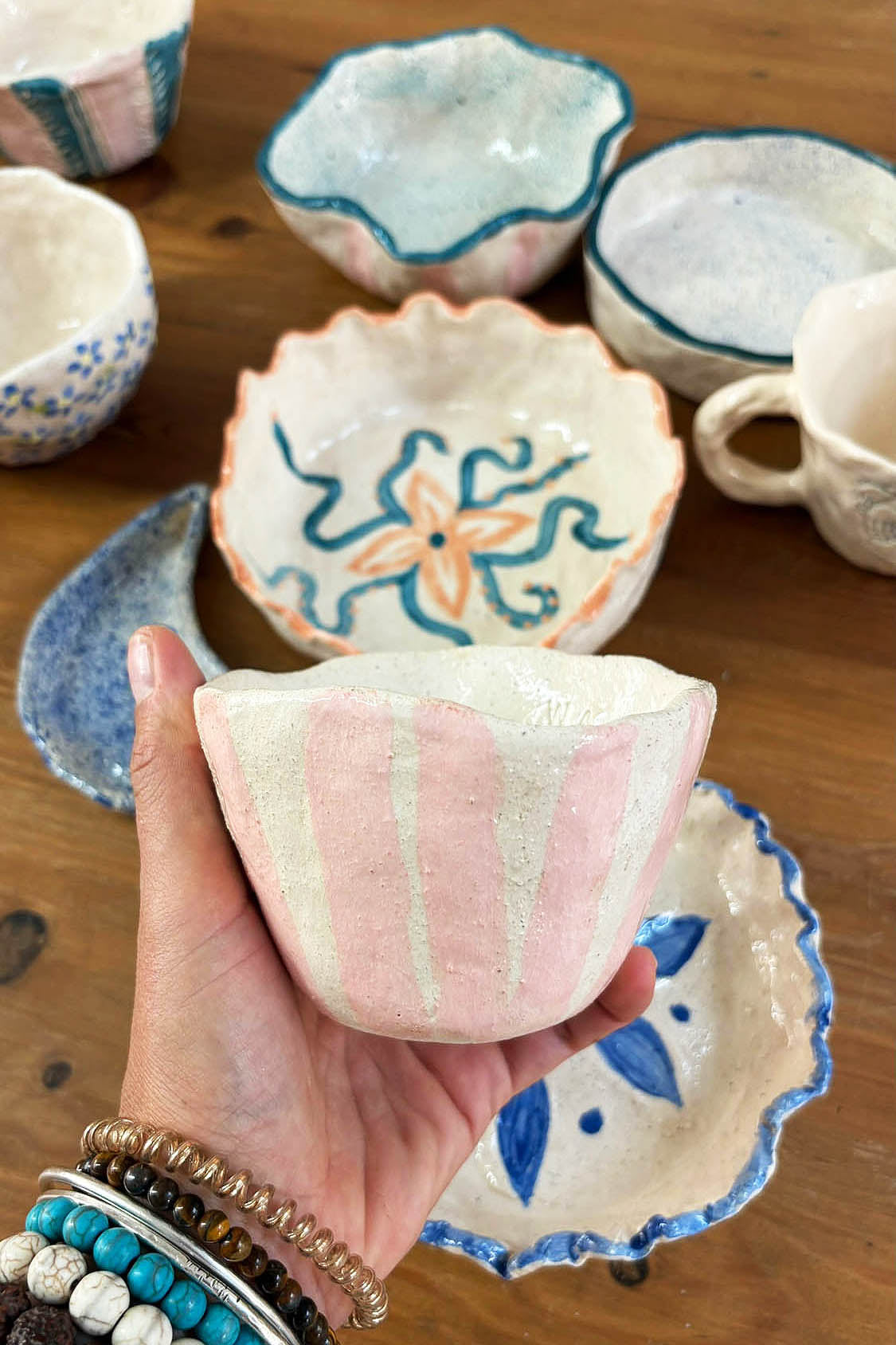 Ceramic Workshop in Lisbon, Portugal. A hand holding a handmade pink and white striped ceramic bowl, with other decorated ceramic dishes arranged on a wooden table in the background.