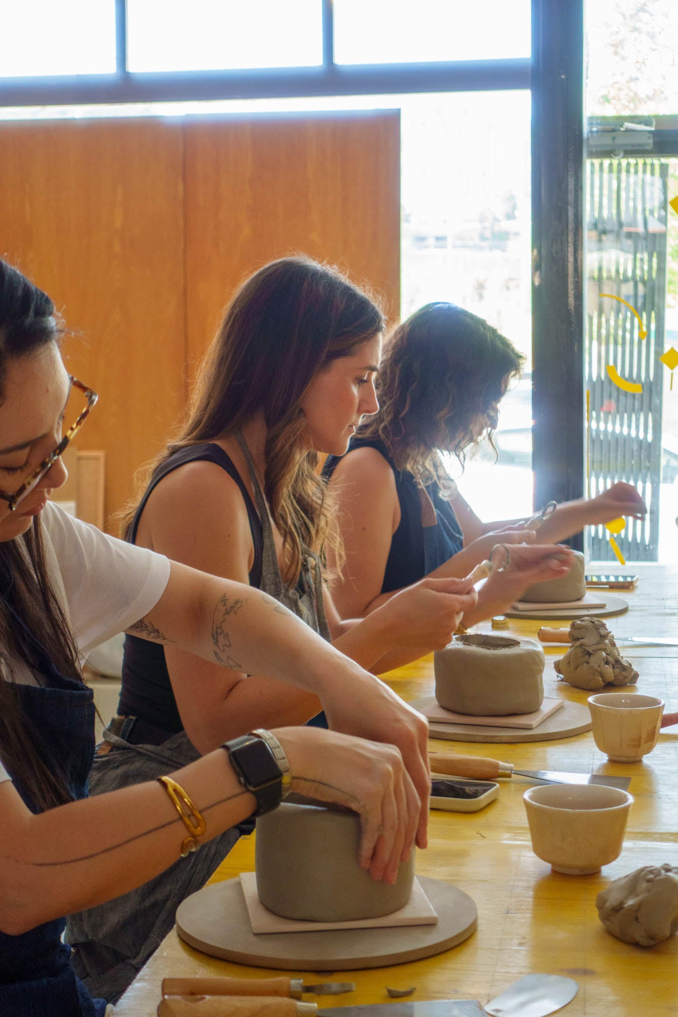 Three women working on pottery at a table with clay pots and tools.