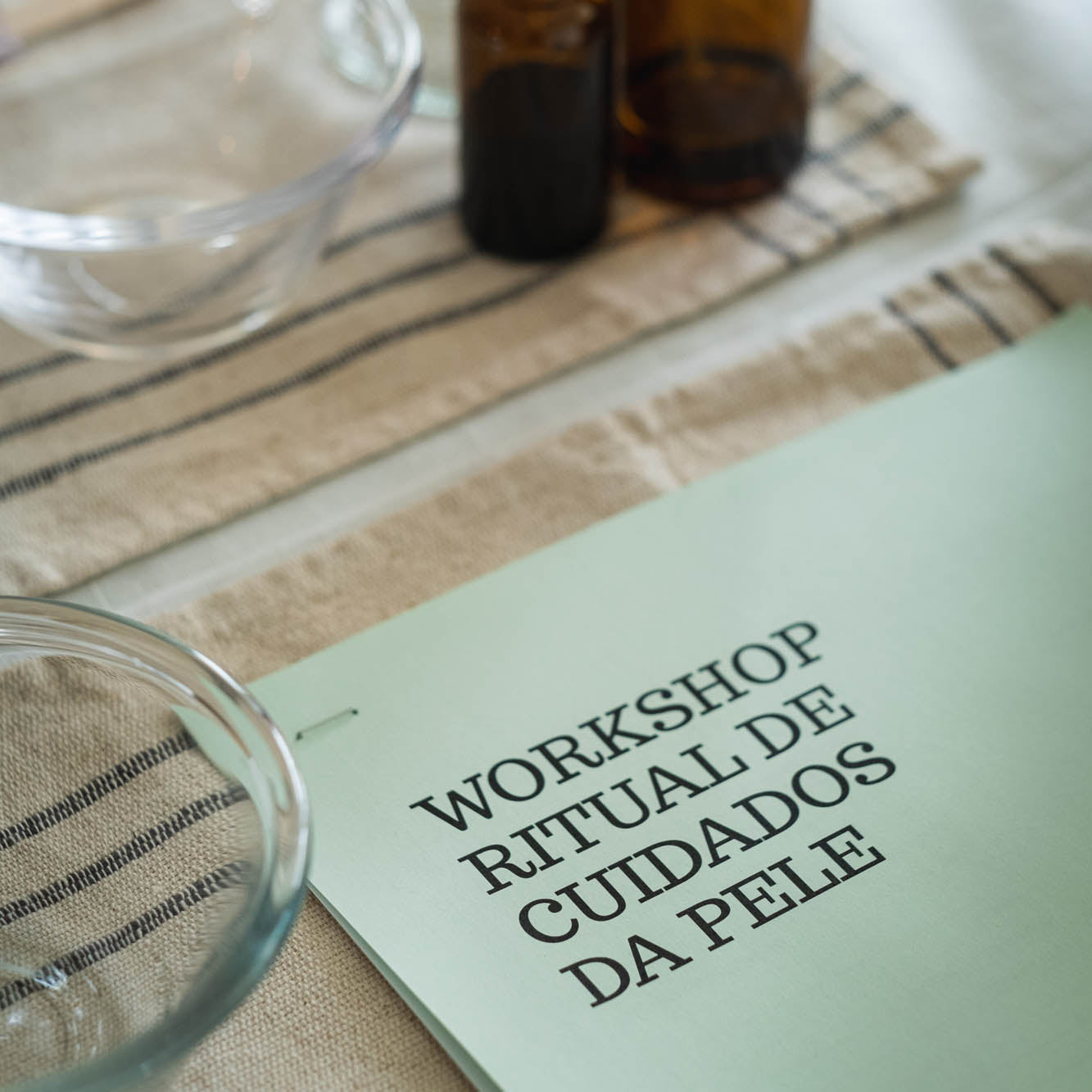 Sheet of paper with 'WORKSHOP RITUAL DE CUIDADOS DA PELE' text on a table with bottles and glasses.