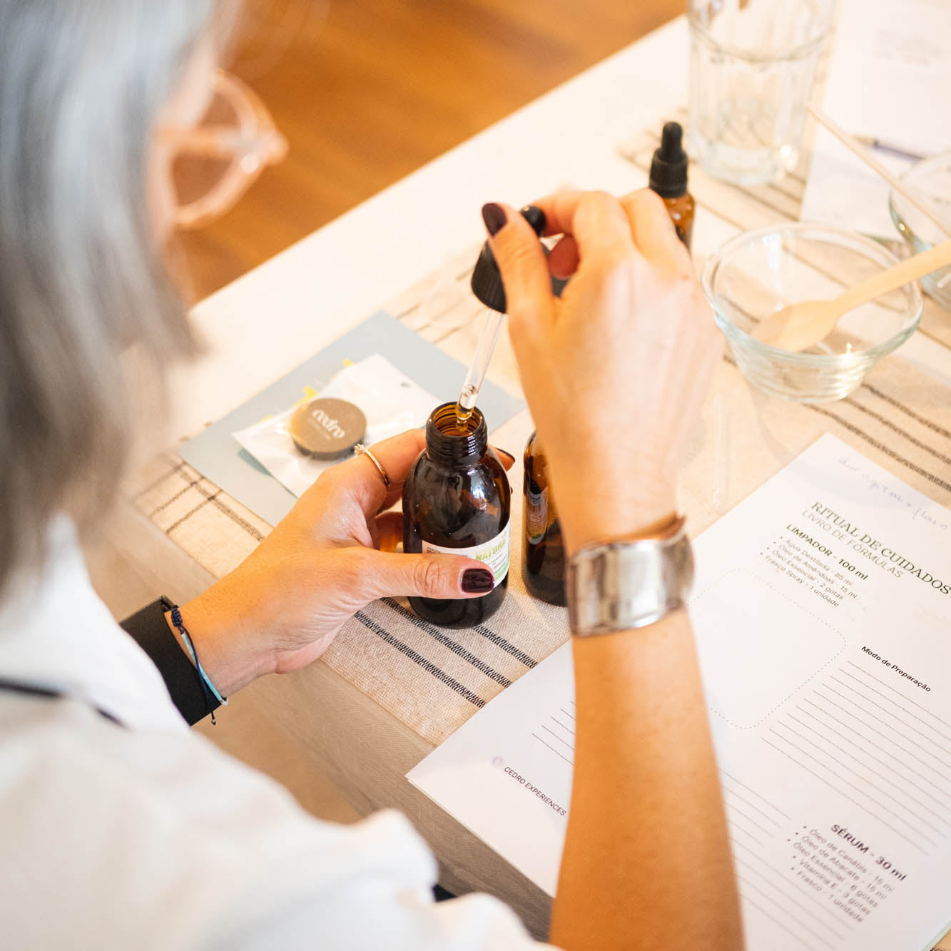 Person using a dropper to apply a liquid from a small bottle on a table with books and a laptop.