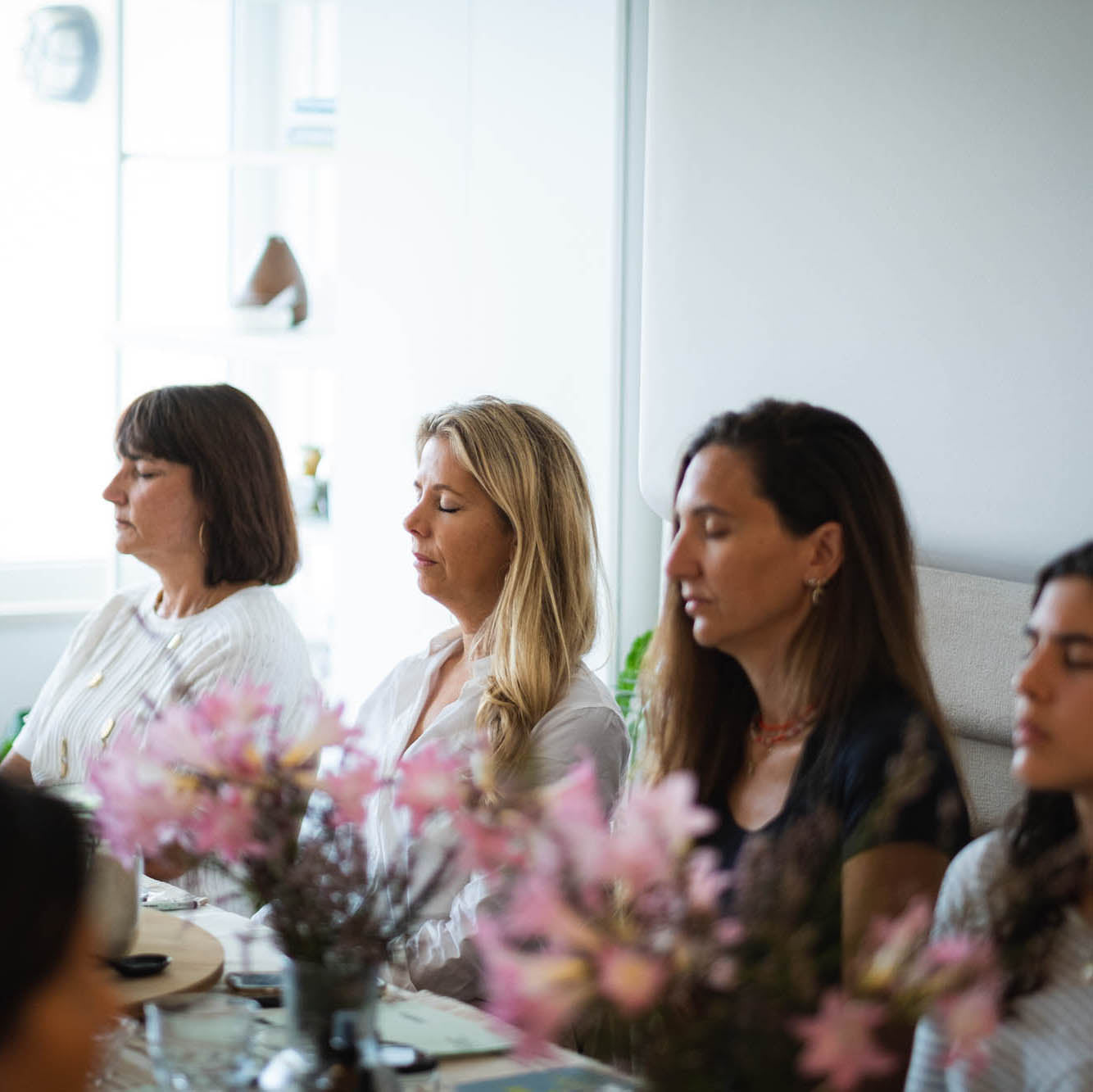Four women sitting together, in a meditation, with a floral arrangement in the foreground.