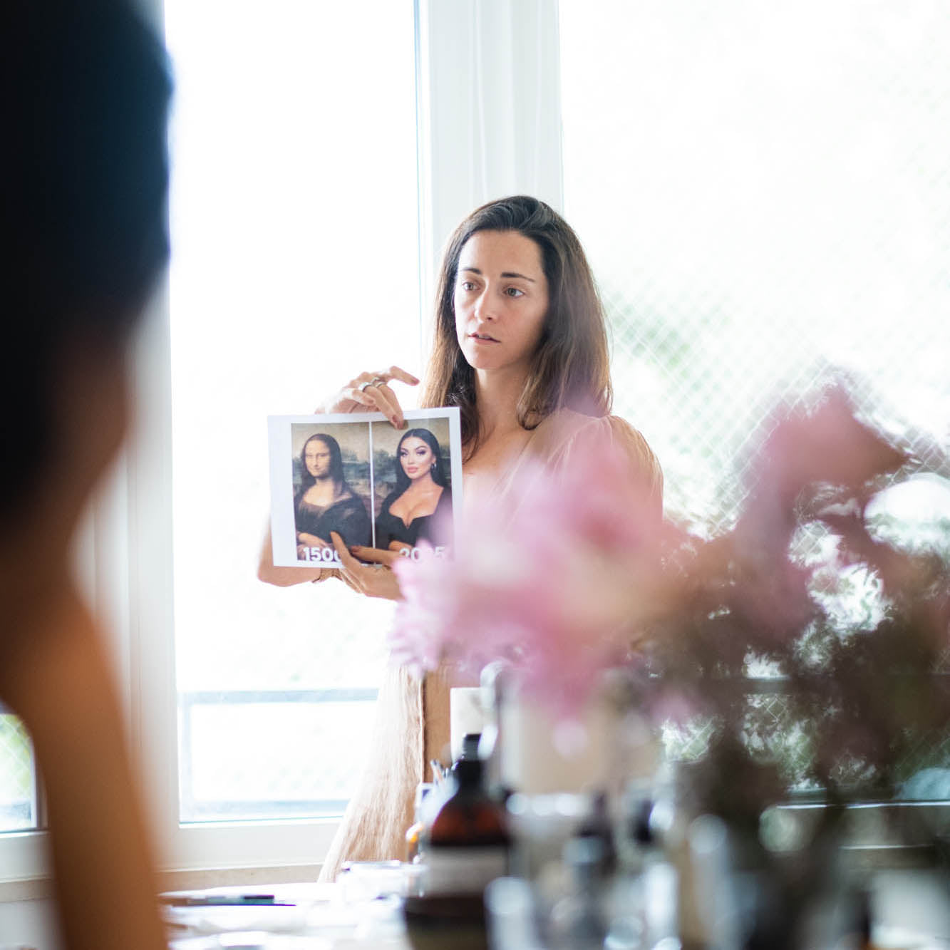 Woman holding a framed picture of a Mona Lisa 1500 and 2025, with a flower arrengement in front of her.