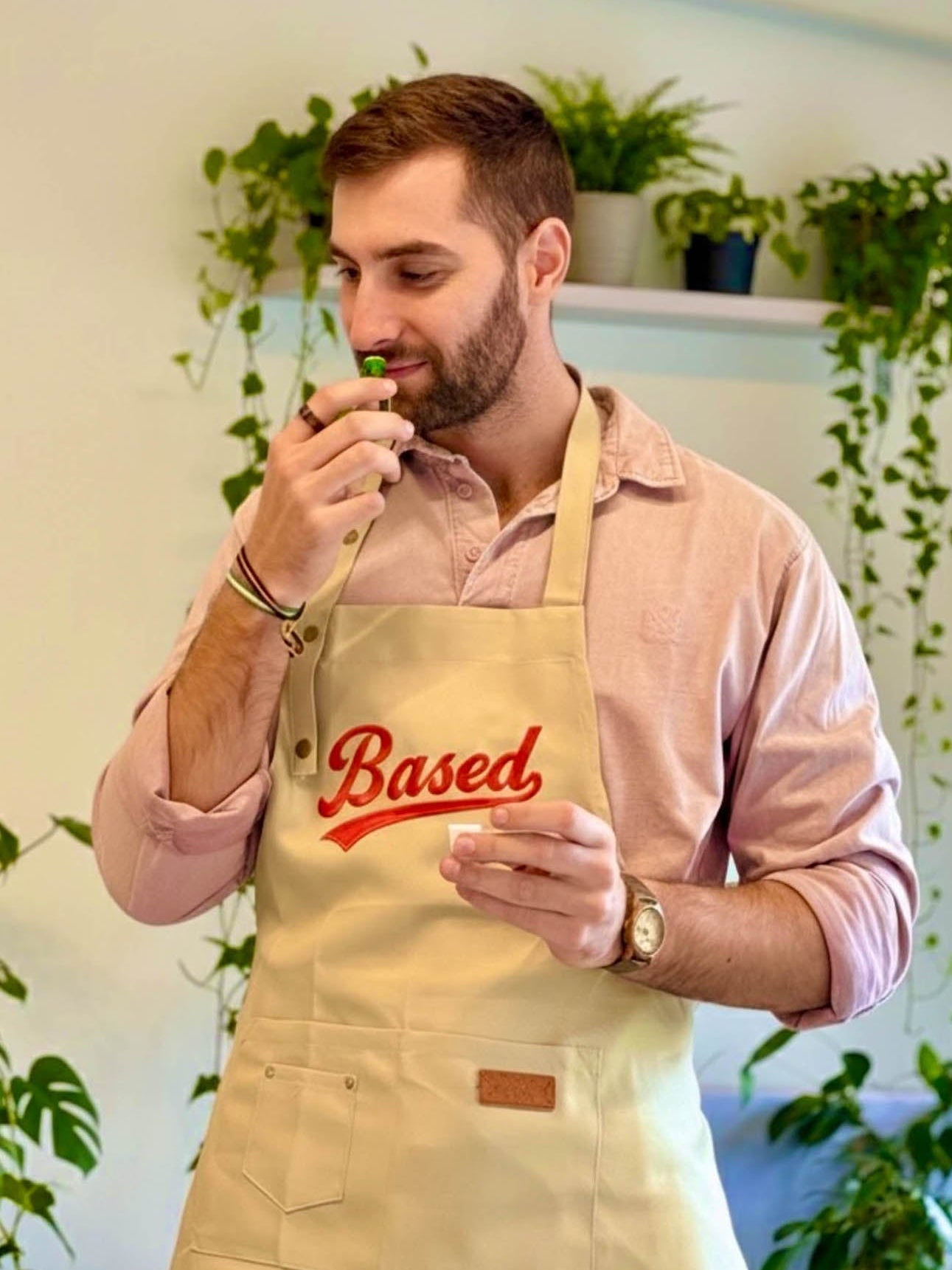 A man wearing a beige apron with 'Based' printed in red script over a pink shirt, holding a small green object near his face in an indoor setting with potted plants.