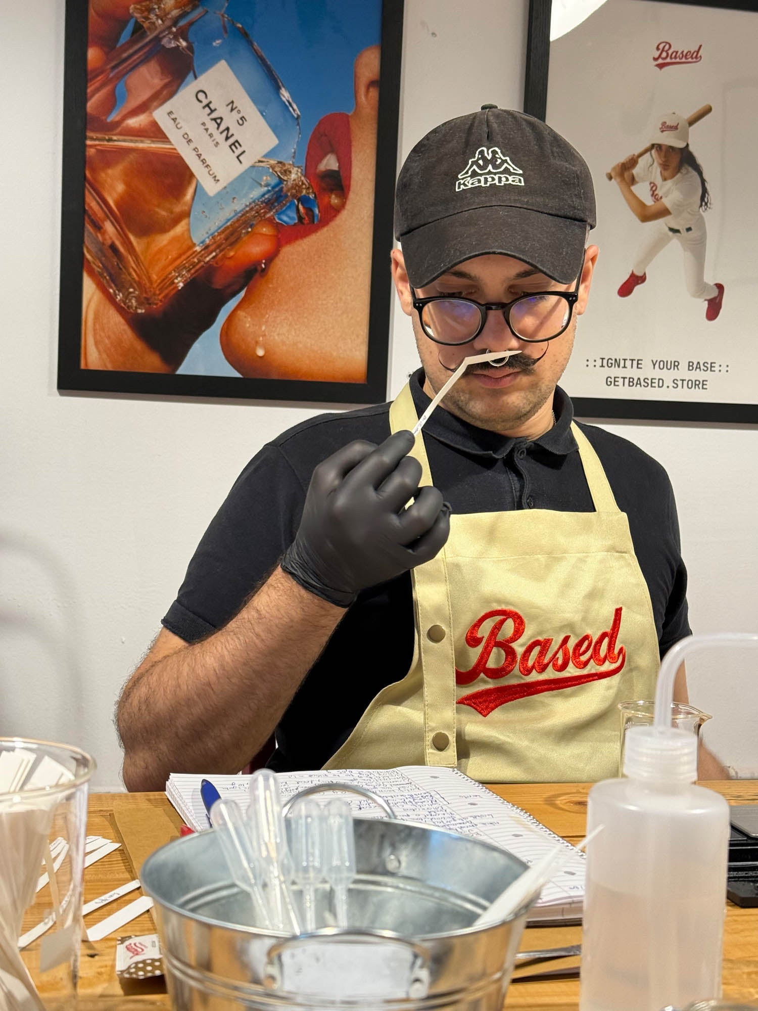 A man, holding a small tool near his mouth while wearing a black glove. He is sitting at a wooden table with a metal bucket, plastic bottles, and papers in a workshop setting with framed art on the wall.